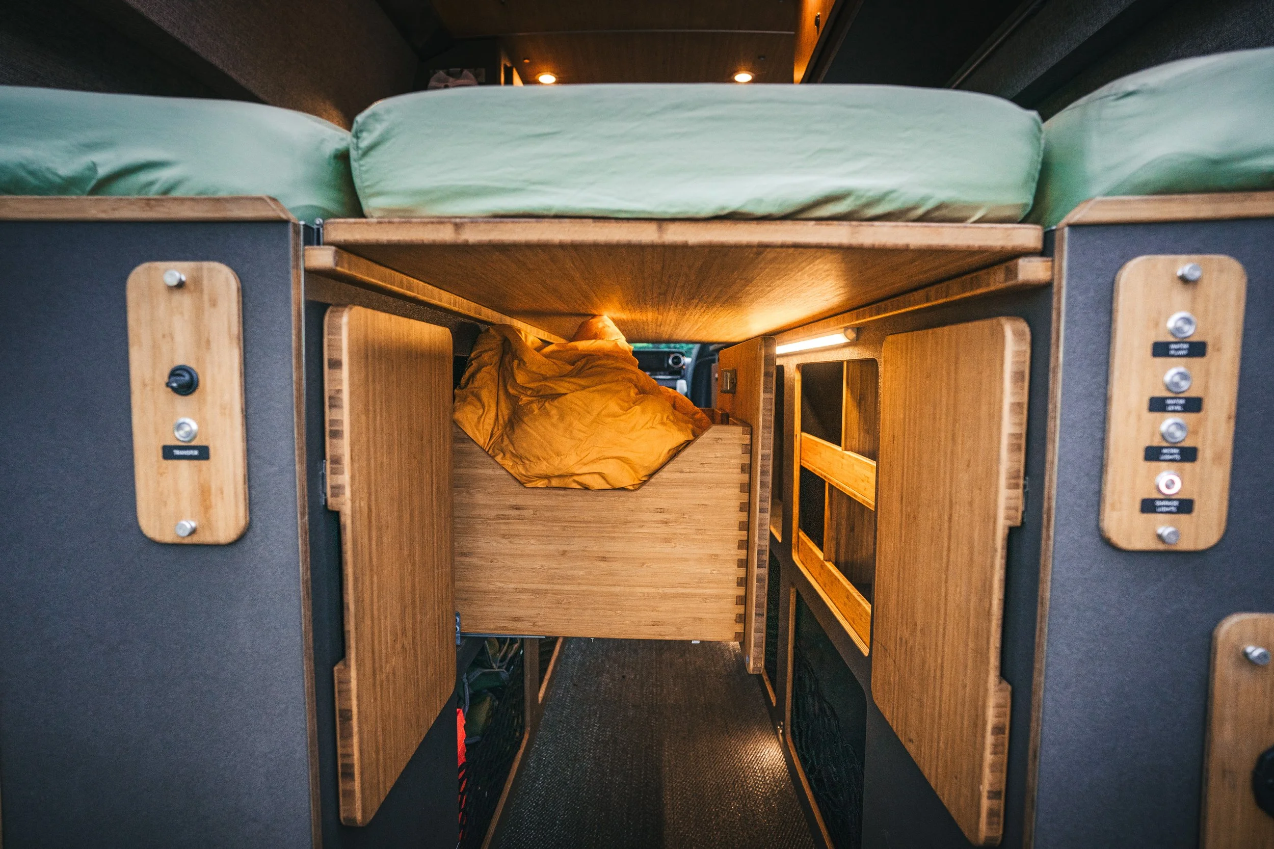 Inside a camper van showing a bed with green bedding above a compact wooden kitchen area.