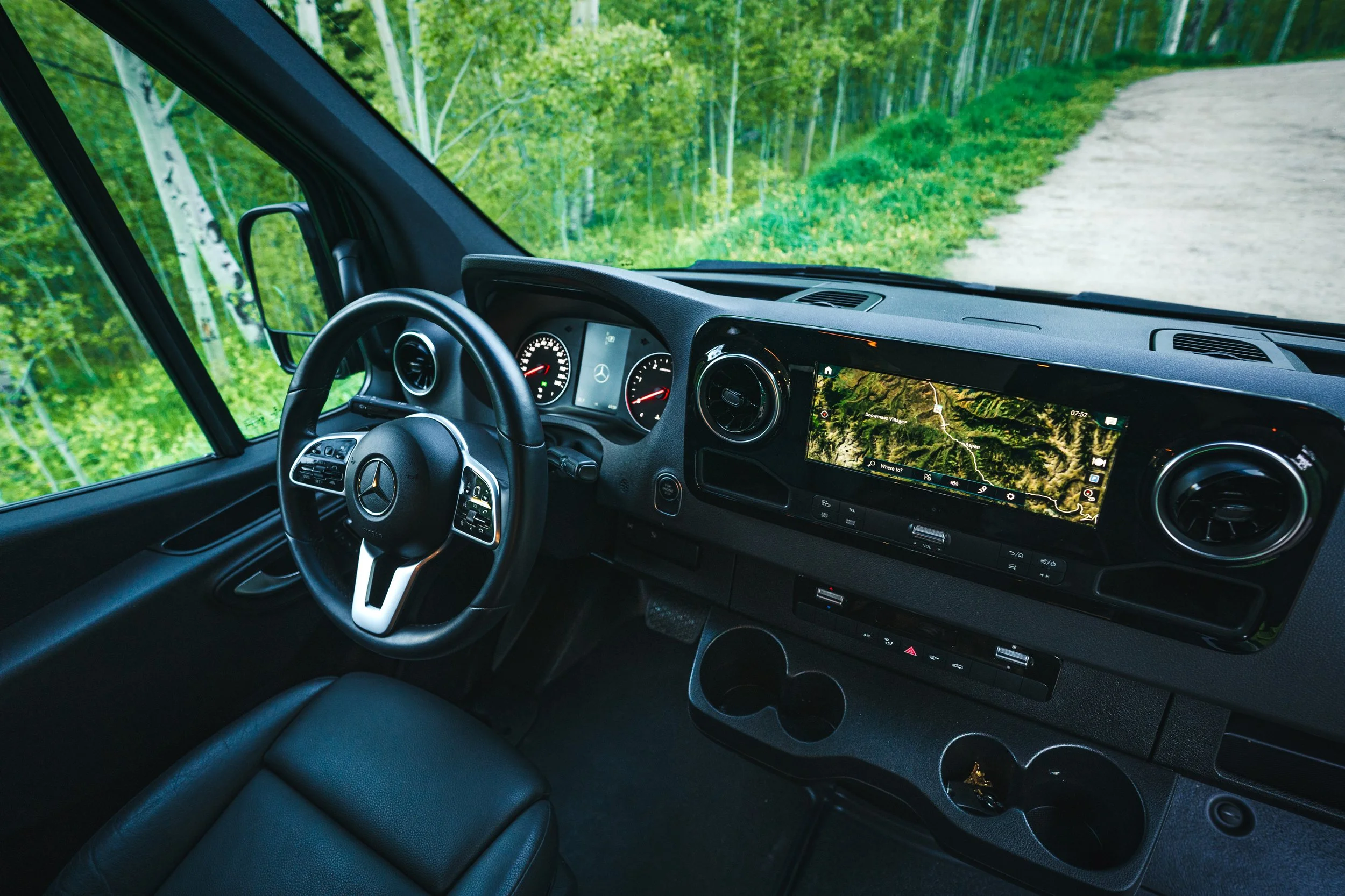 Interior of a Mercedes-Benz van showing steering wheel, dashboard with navigation screen, and a view of a lush green forest outside through the windows.