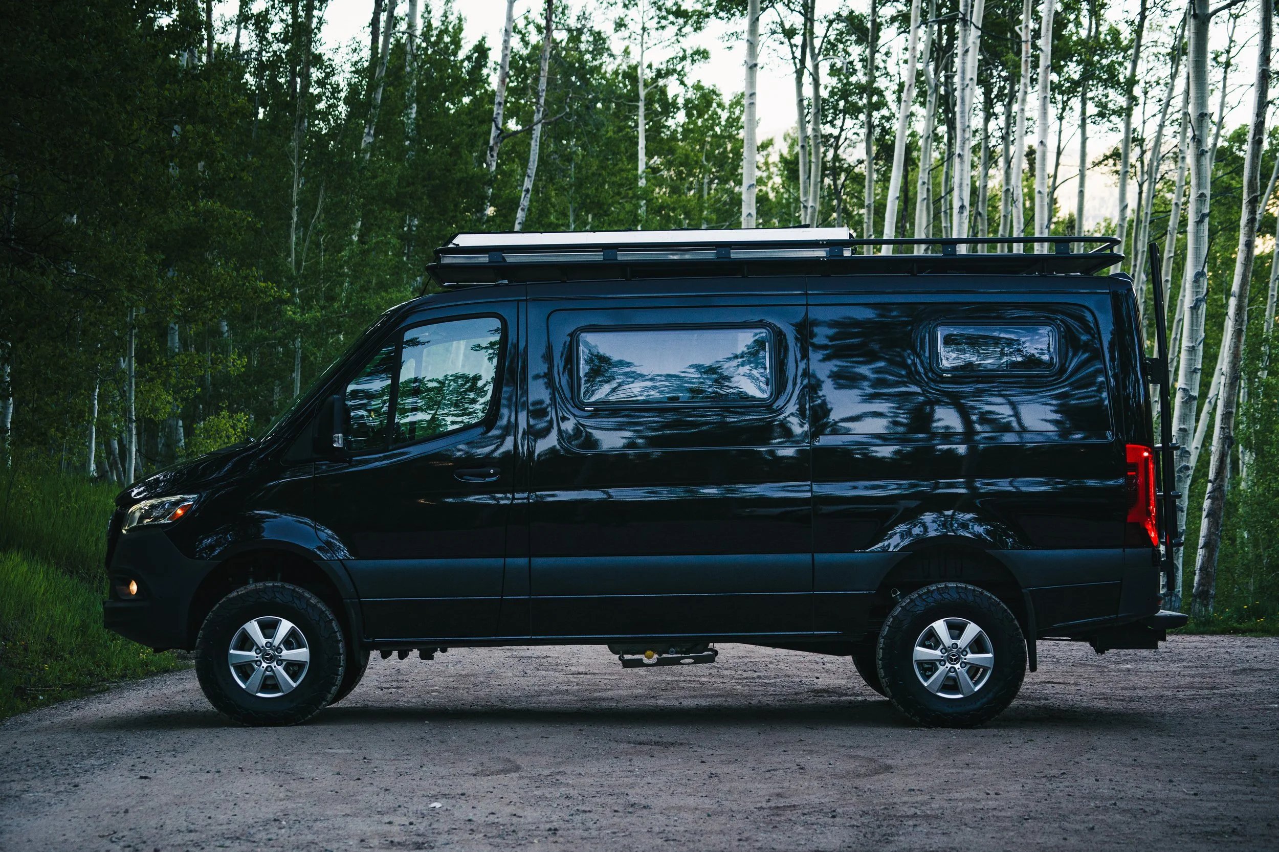 Black camper van parked on a dirt road in a forest with green trees and white birch trees in the background.