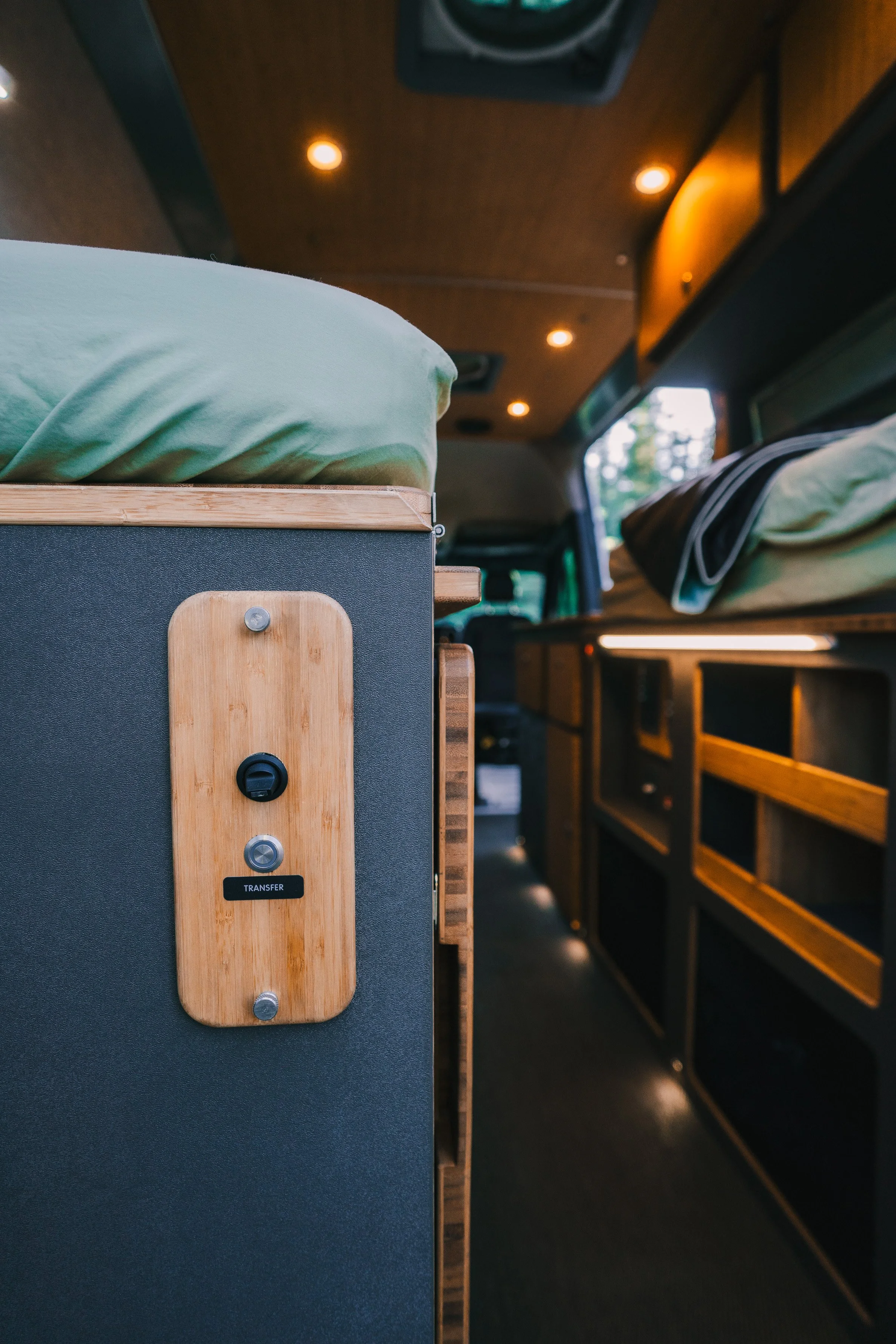 Interior of a vehicle or camper with built-in beds, wooden cabinetry, and lighting, focusing on a power outlet and transfer switch on a side panel.