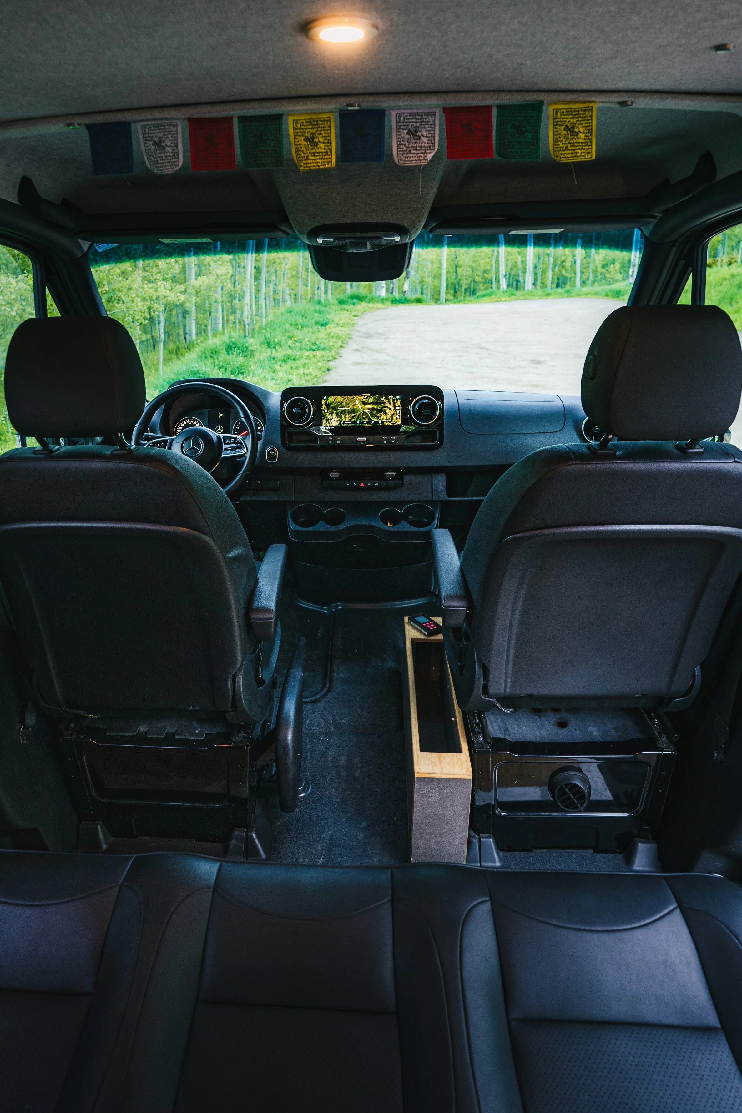 Interior view of a vehicle showing the front seats, dashboard, steering wheel, and windshield looking out to a grassy path with trees in the background.