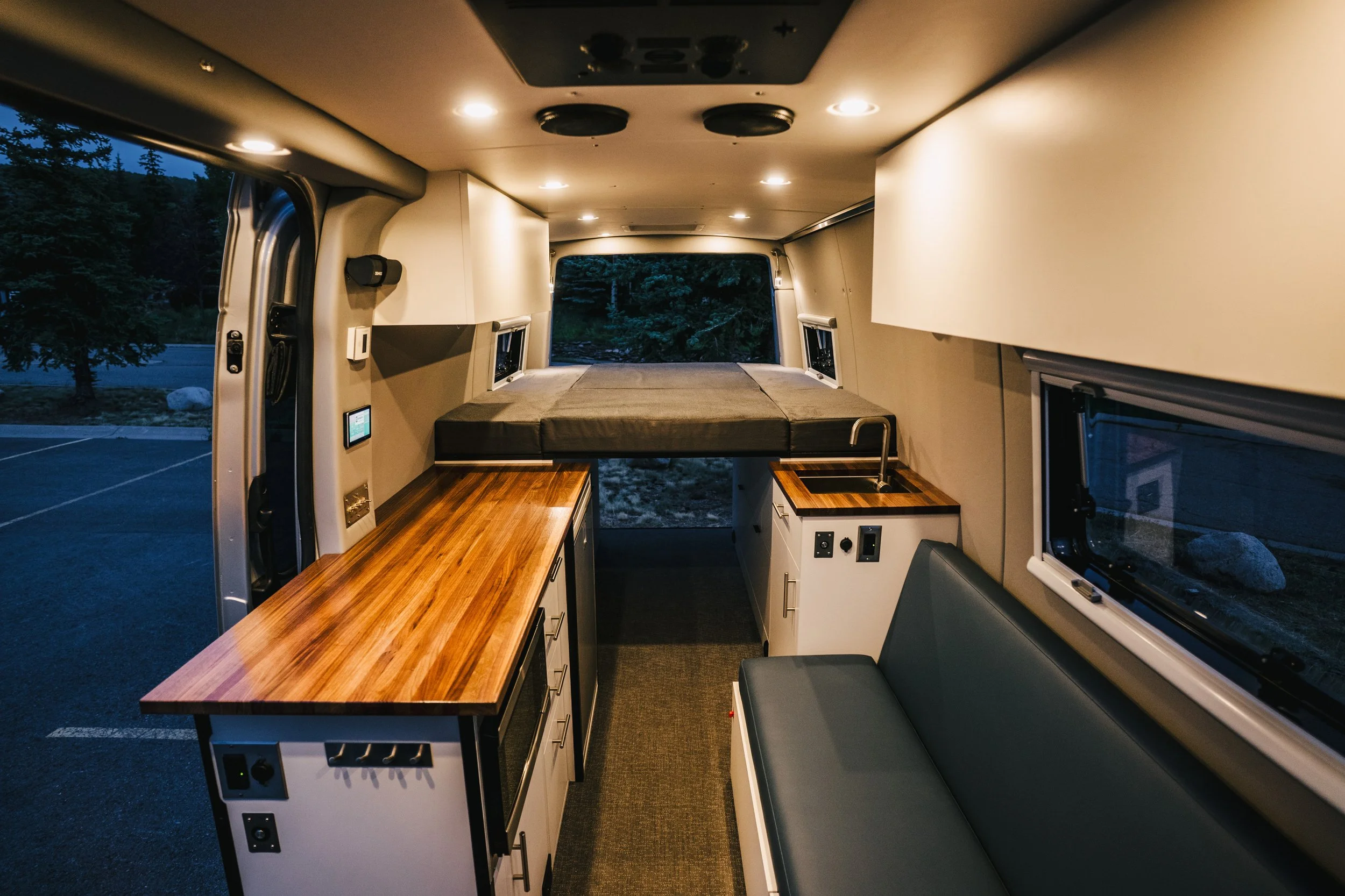 Interior of a converted van with a bed at the rear, wooden countertops on the sides with a sink, small appliances, and a seating area along the right side window, illuminated by ceiling lights.
