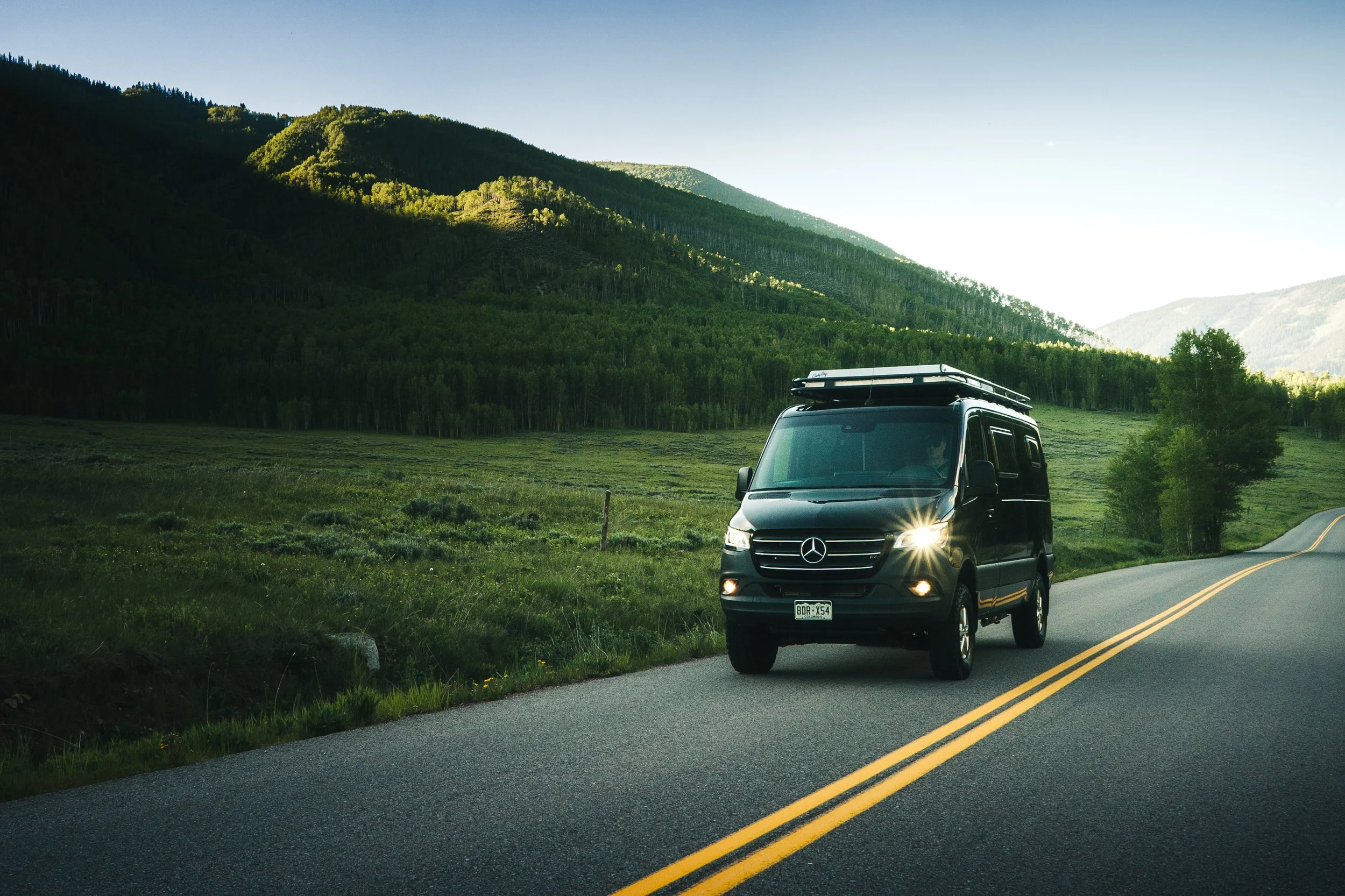 Black Mercedes-Benz van driving on a winding road through green countryside with mountains in the background, in bright daylight.