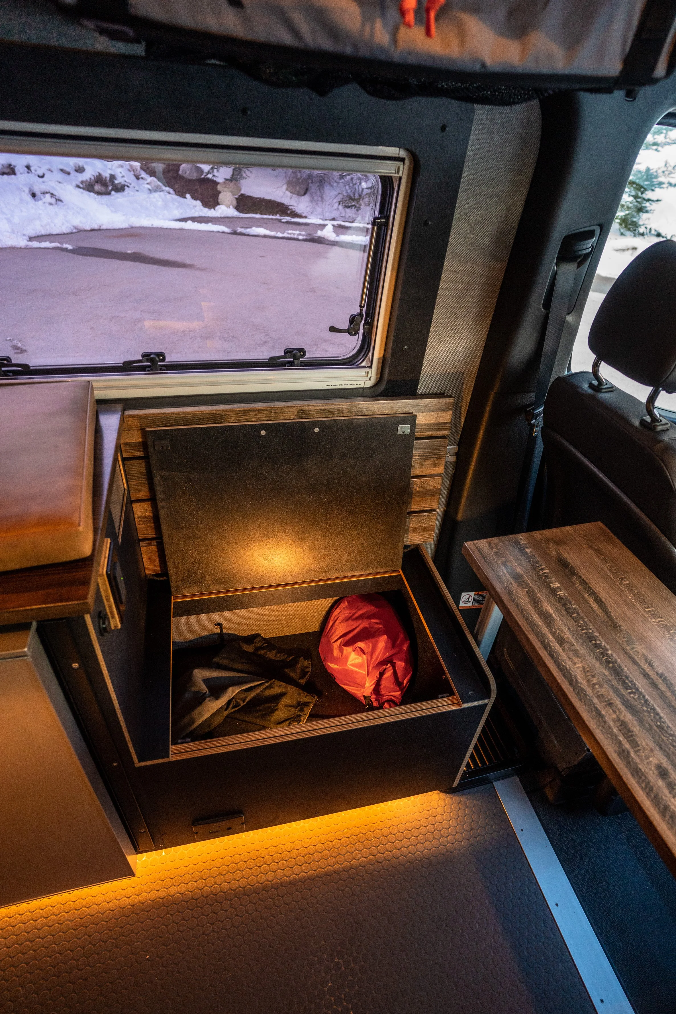A storage compartment inside a vehicle, with luggage and orange lighting at the bottom, near a window showing snow outside.
