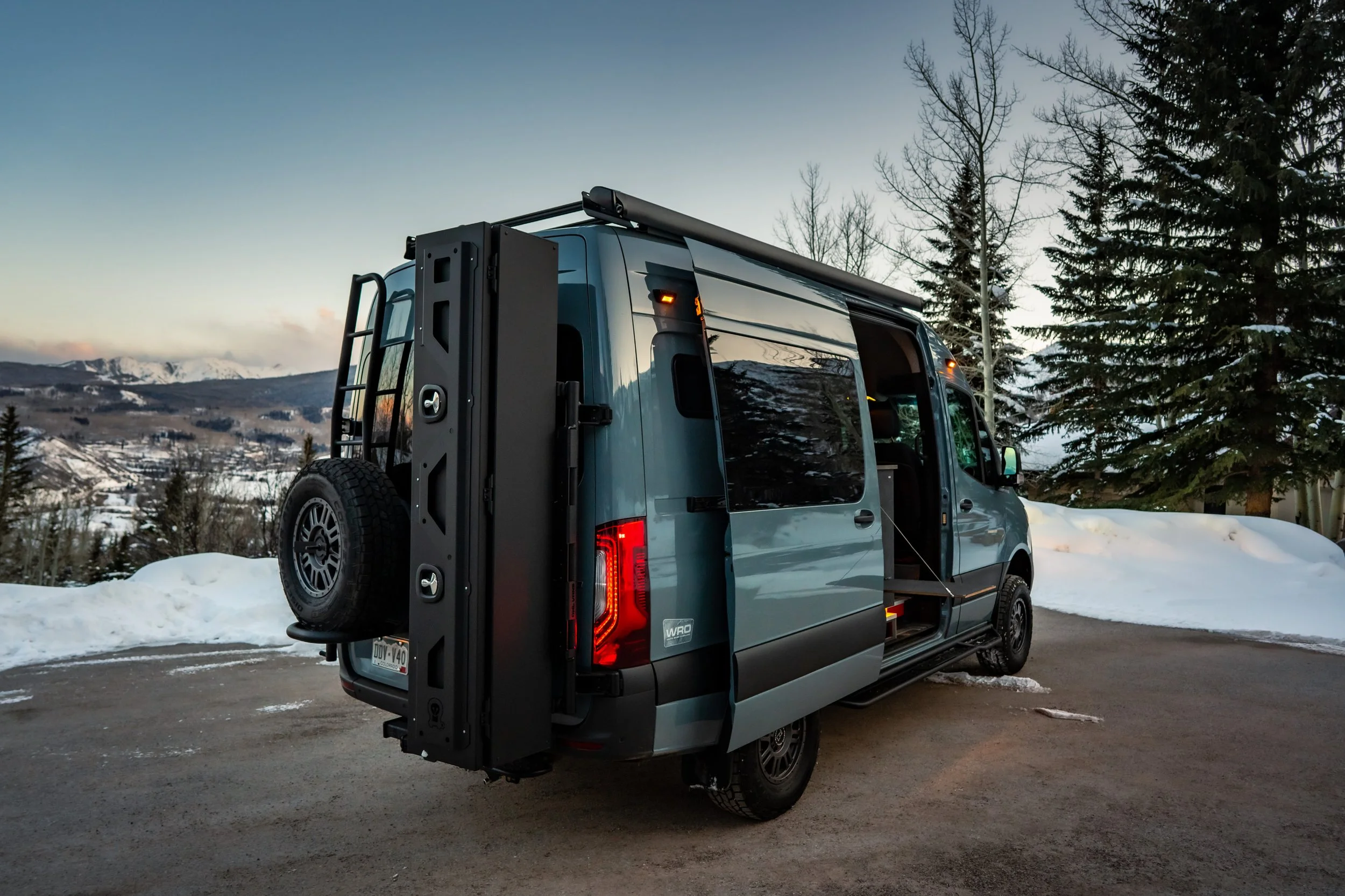 A blue camper van parked outdoors in winter with snow on the ground and pine trees in the background, mountain range in the distance, and a partly cloudy sky.