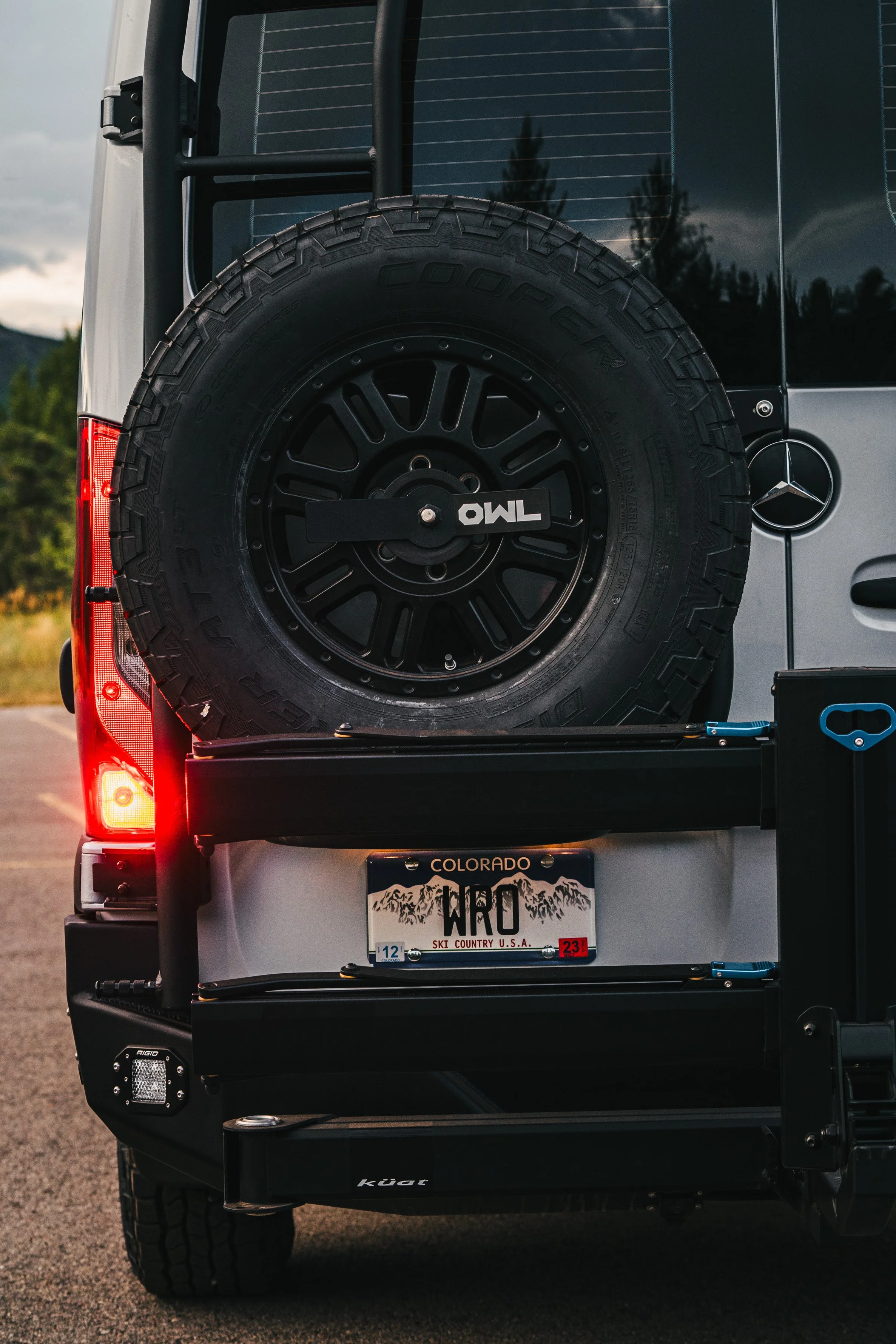 Rear view of a Mercedes-Benz vehicle with a spare tire mounted on the back, a Colorado license plate with mountains and the word 'WRO', and red tail lights illuminated, parked outdoors.