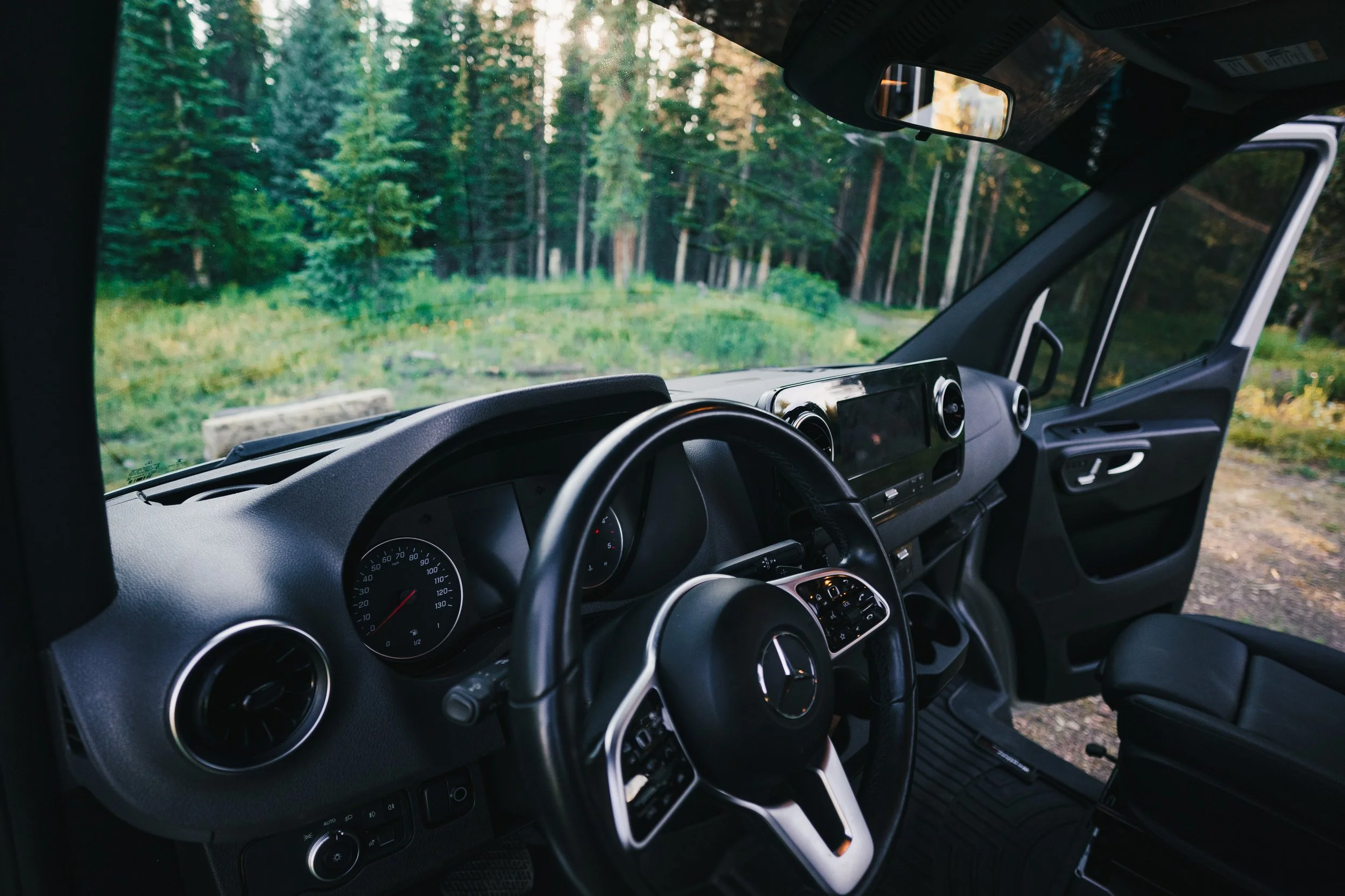 Interior of a vehicle with a Mercedes-Benz steering wheel, dashboard, and view of a forested area outside the windshield.