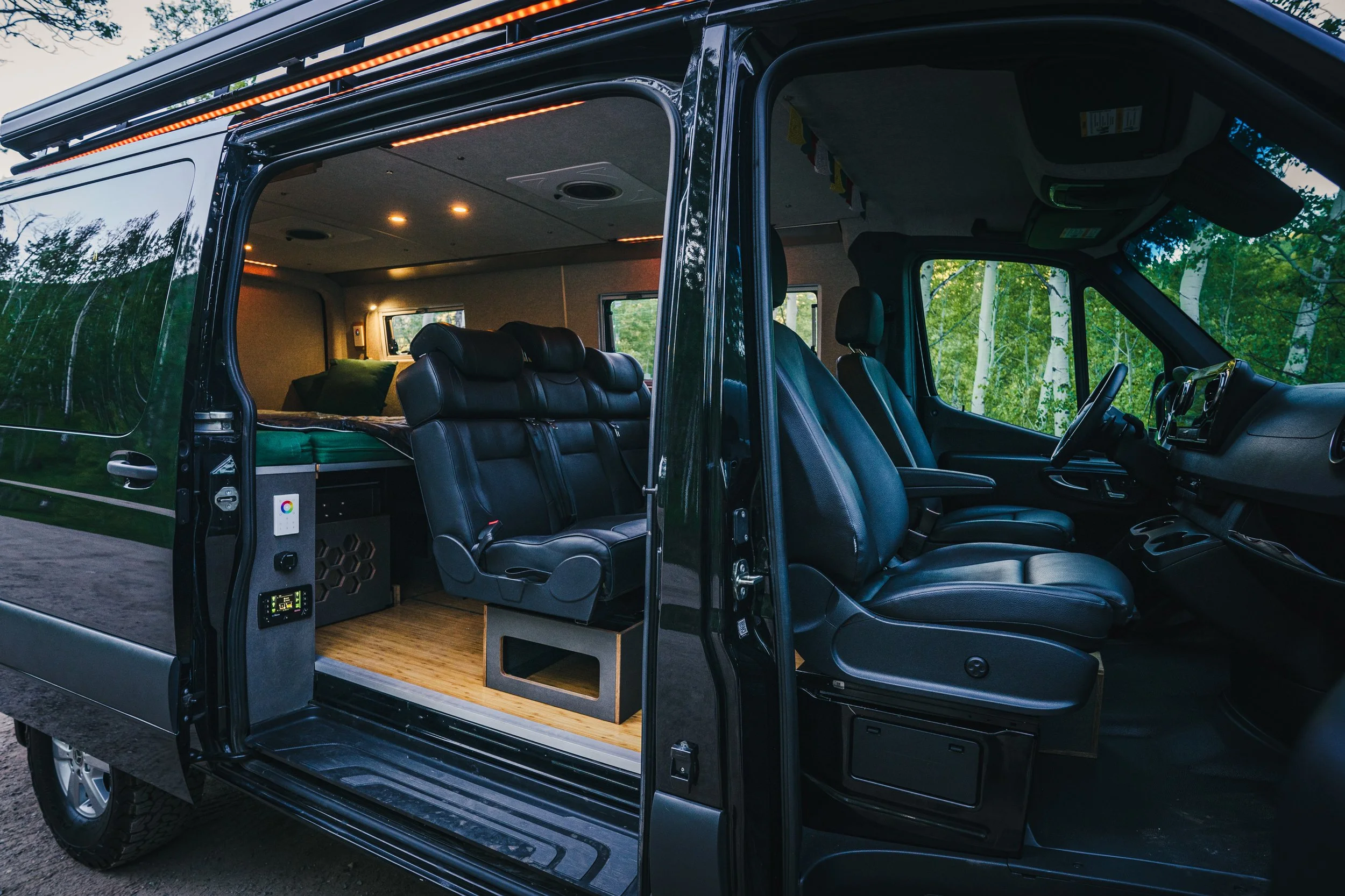 Interior view of a converted van showing black leather seats, a bed in the back, and a decorated ceiling with string lights, situated in a green wooded area.