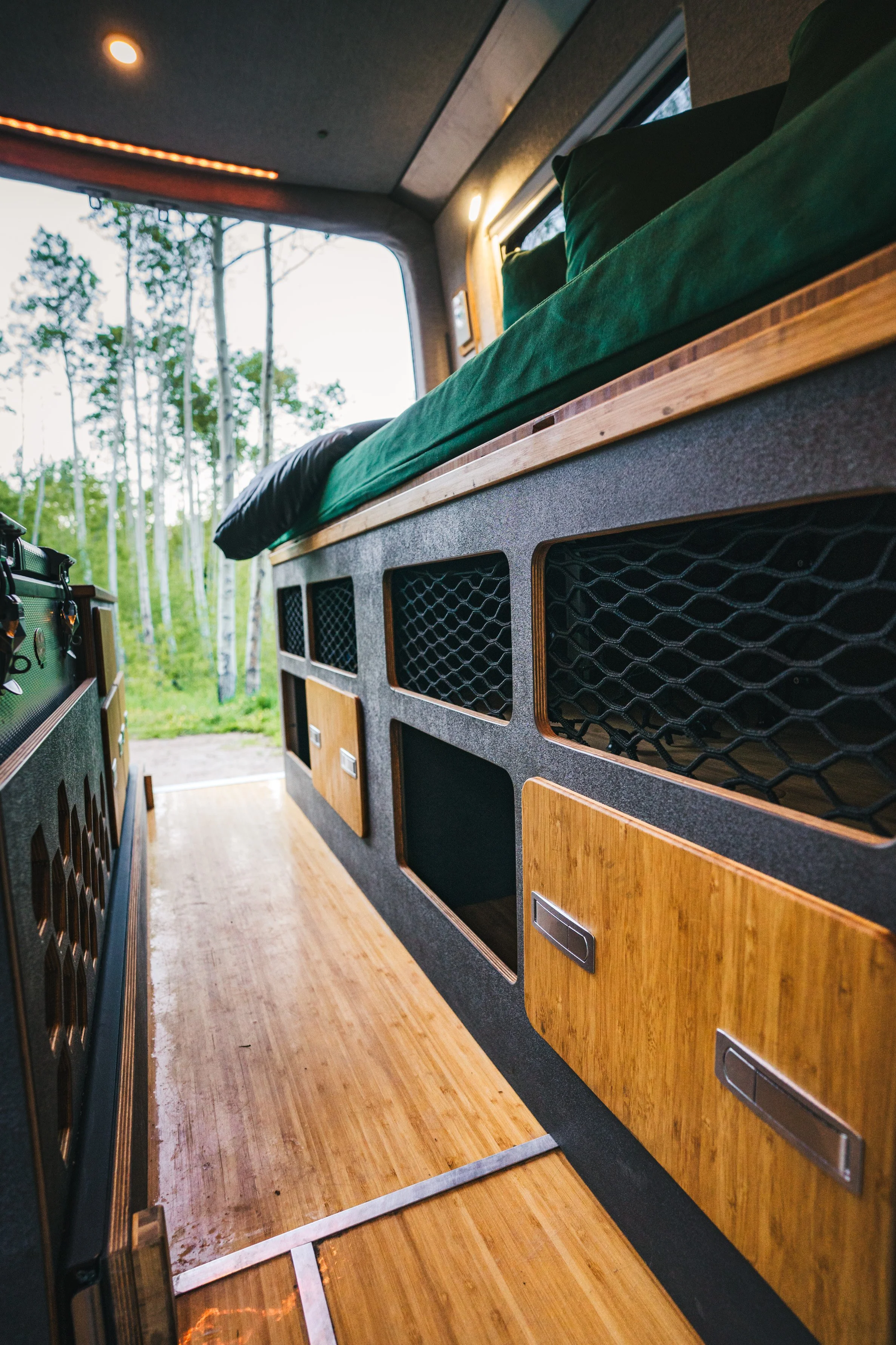 Interior view of a camper or van with wooden storage compartments and a bed with green bedding, set against a backdrop of trees outside the open door.