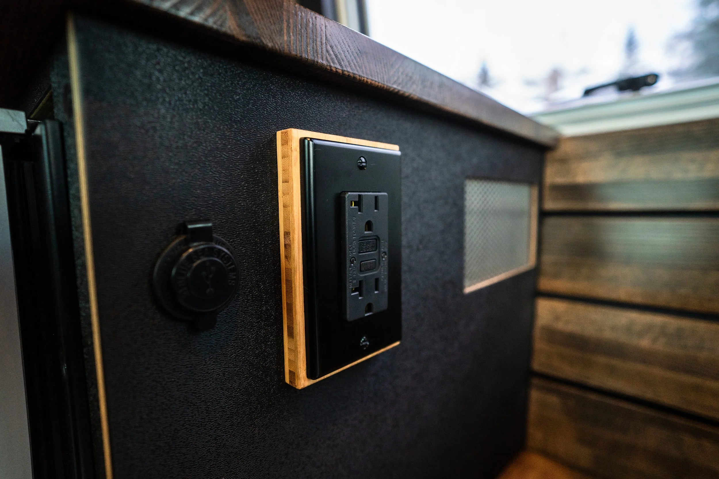 Close-up of a black electrical outlet with a wooden border on a dark textured wall, with a view of a window and a blurred outdoor scene in the background.