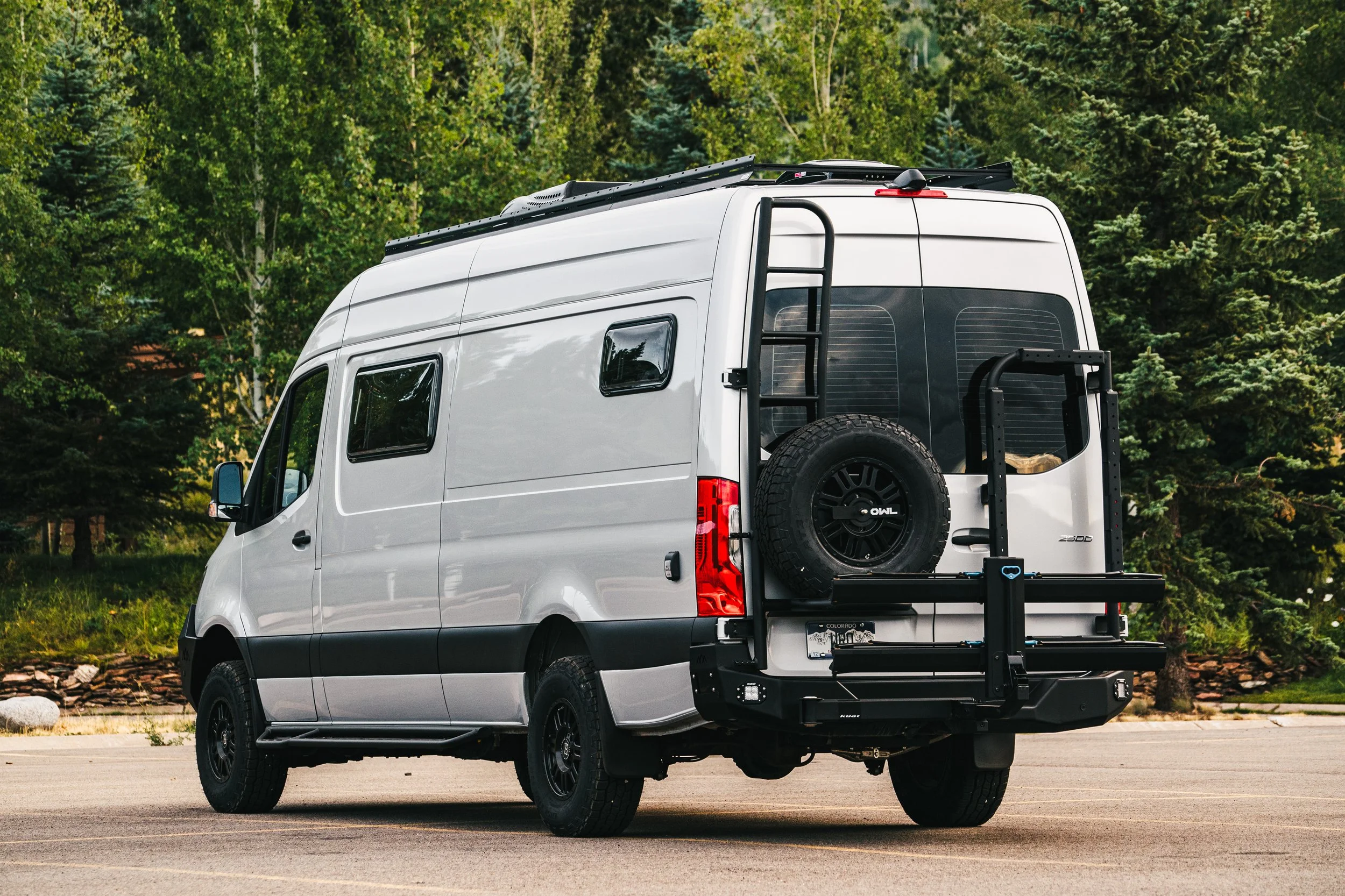 Silver Sprinter camper van parked on a paved surface with green trees in the background, featuring a spare tire mounted on the back and a rear bike rack.