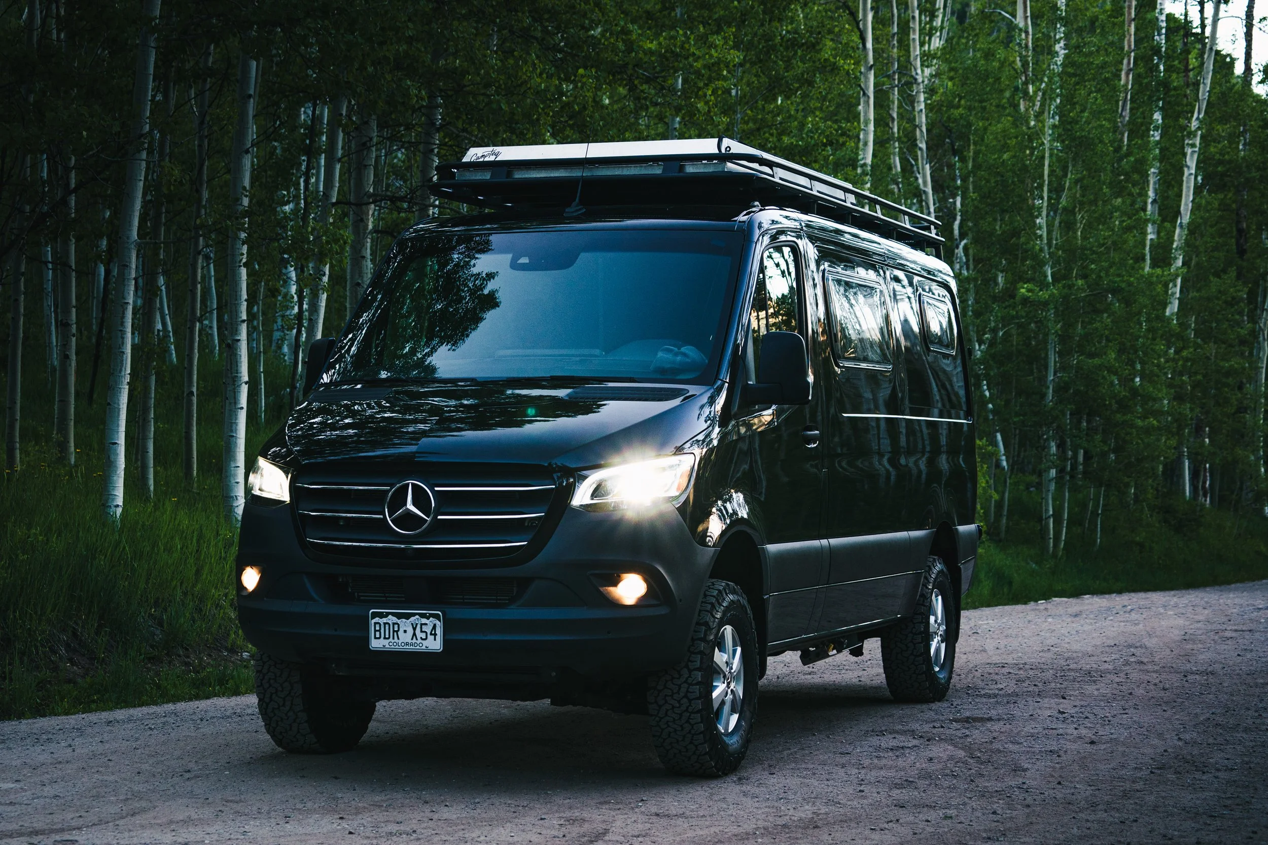 Black Mercedes-Benz camper van with roof rack driving on dirt road surrounded by green trees