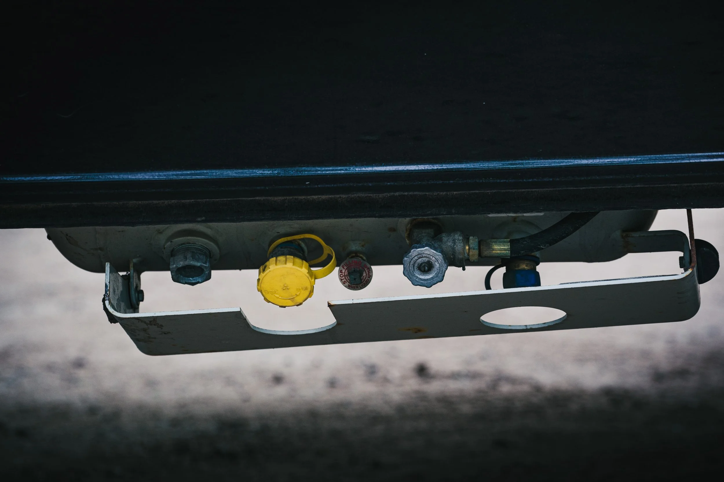 Close-up of a metal box with gas fittings and valves under a black surface, including a yellow valve handle and a red warning label.