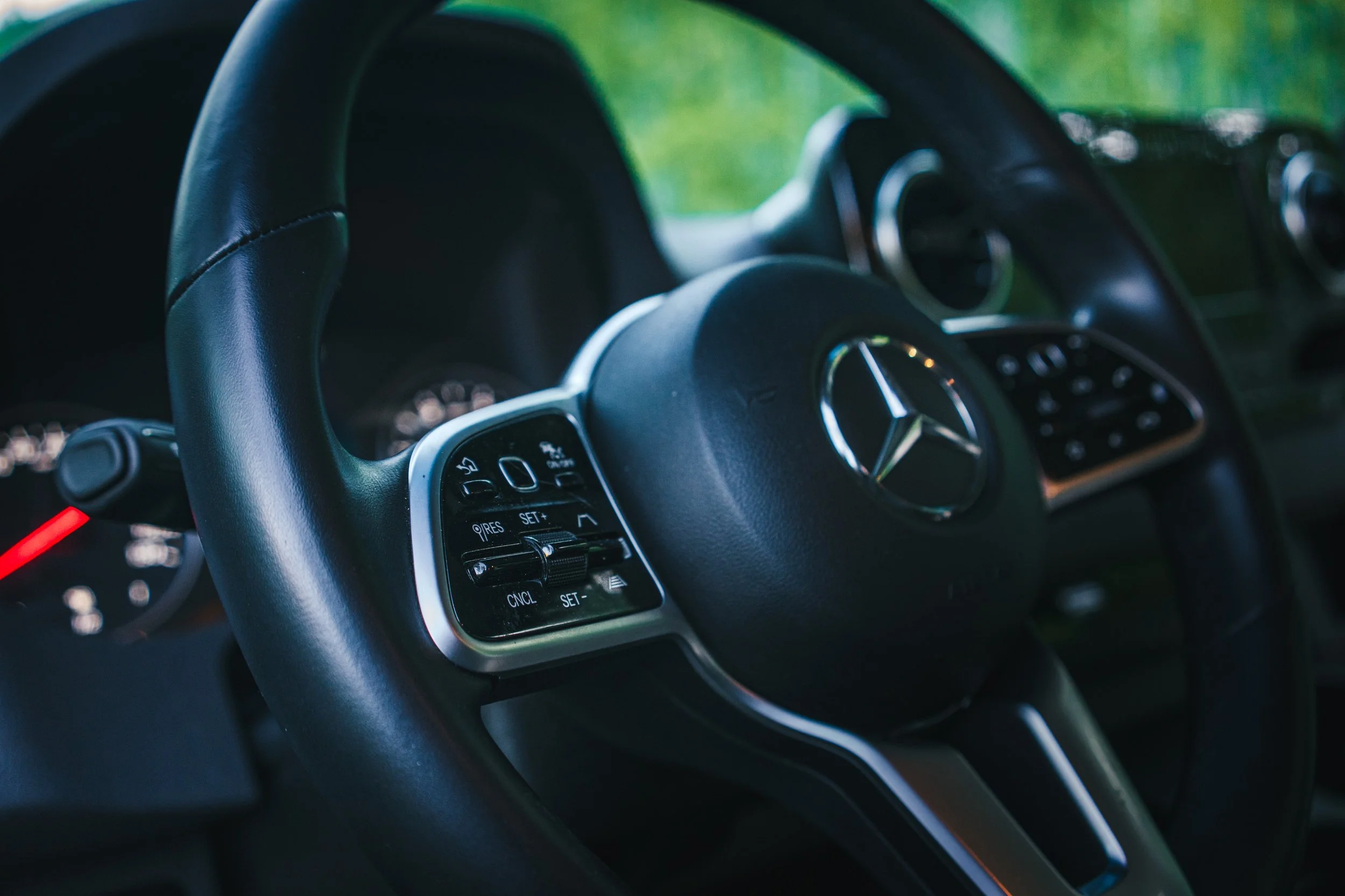 Close-up of a black Mercedes-Benz steering wheel with control buttons, dashboard, and tachometer visible in the background.