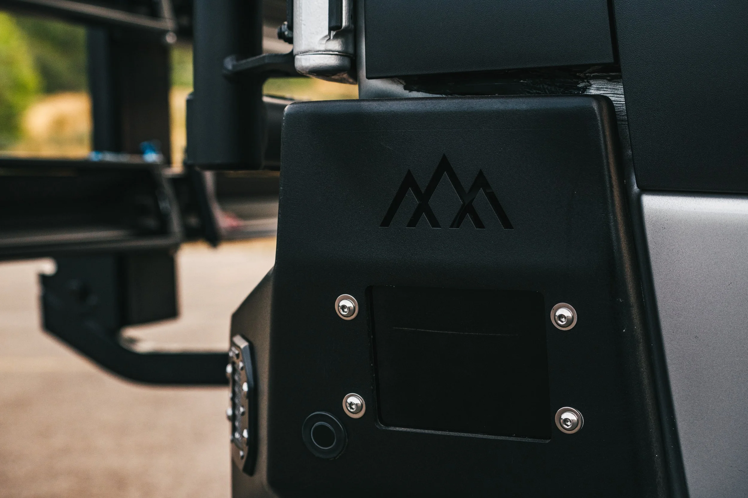 Close-up of a black piece of outdoor equipment with a mountain-like logo cutout and a small circular button or sensor on the lower left, with blurred outdoor background.