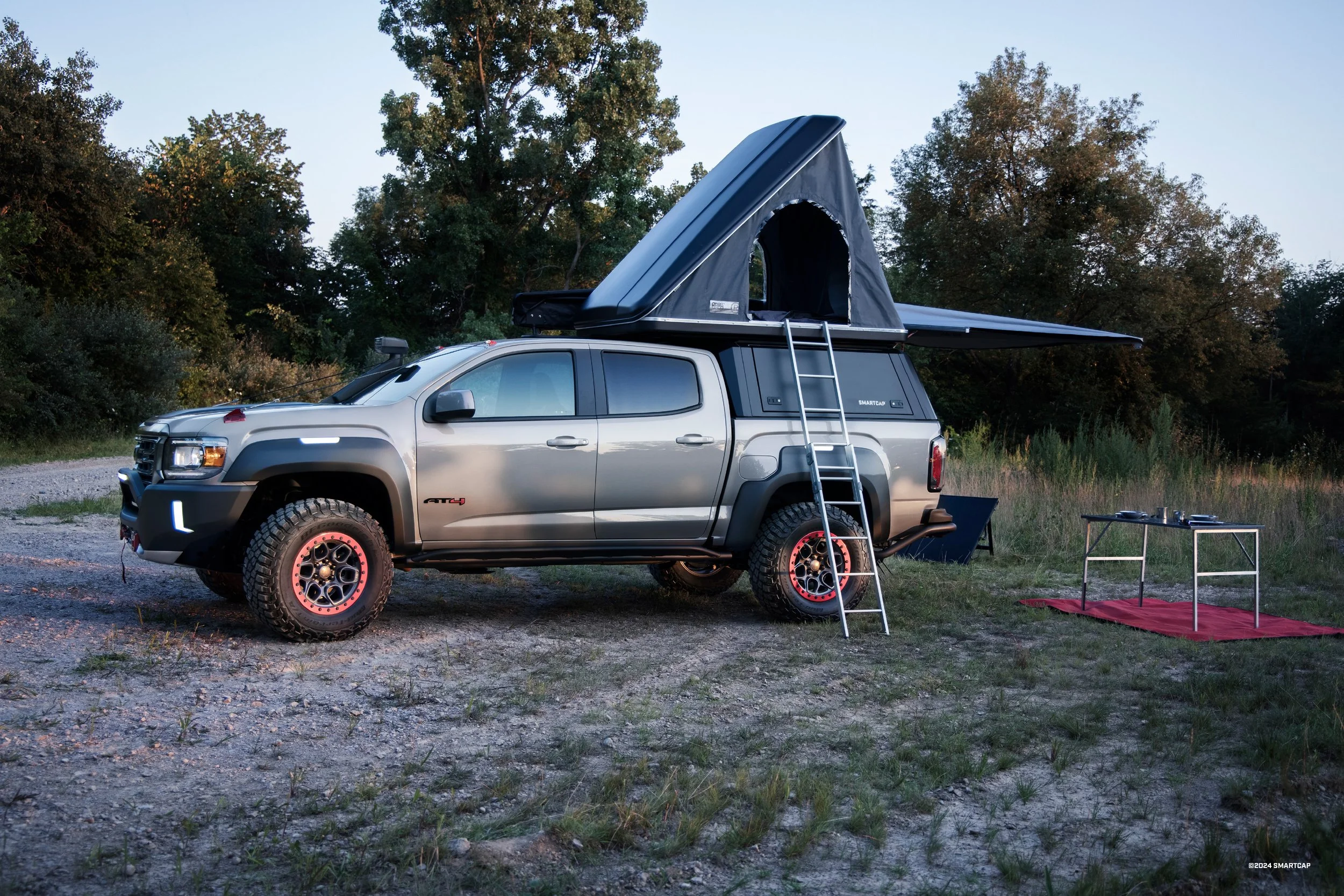 A silver pickup truck with a rooftop pop-up tent set up on a grassy and dirt area, surrounded by trees, with outdoor camping tables and chairs nearby during dusk.