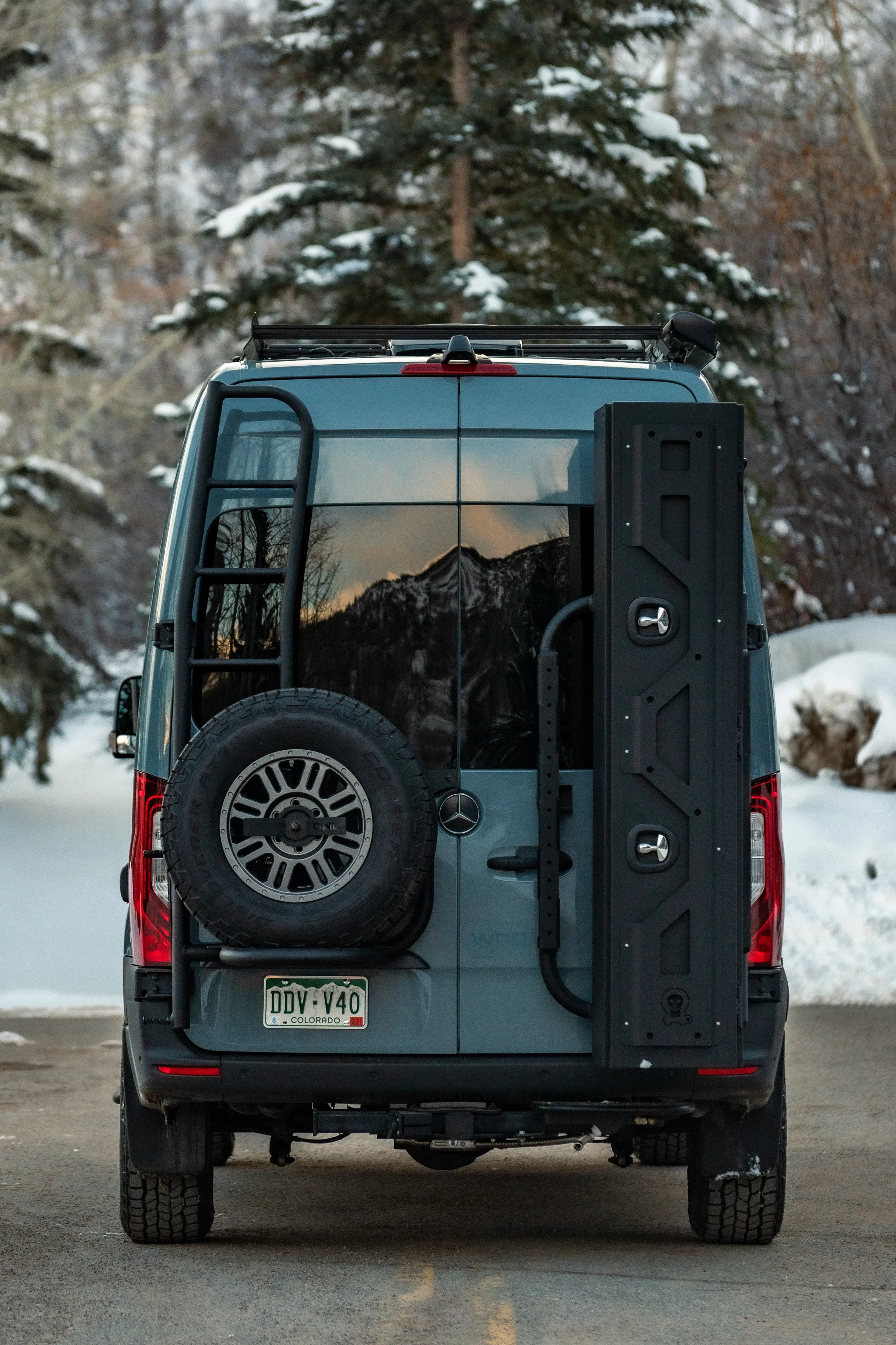 Back of a blue Mercedes-Benz van in a snowy outdoor setting with mountains and trees in the background.