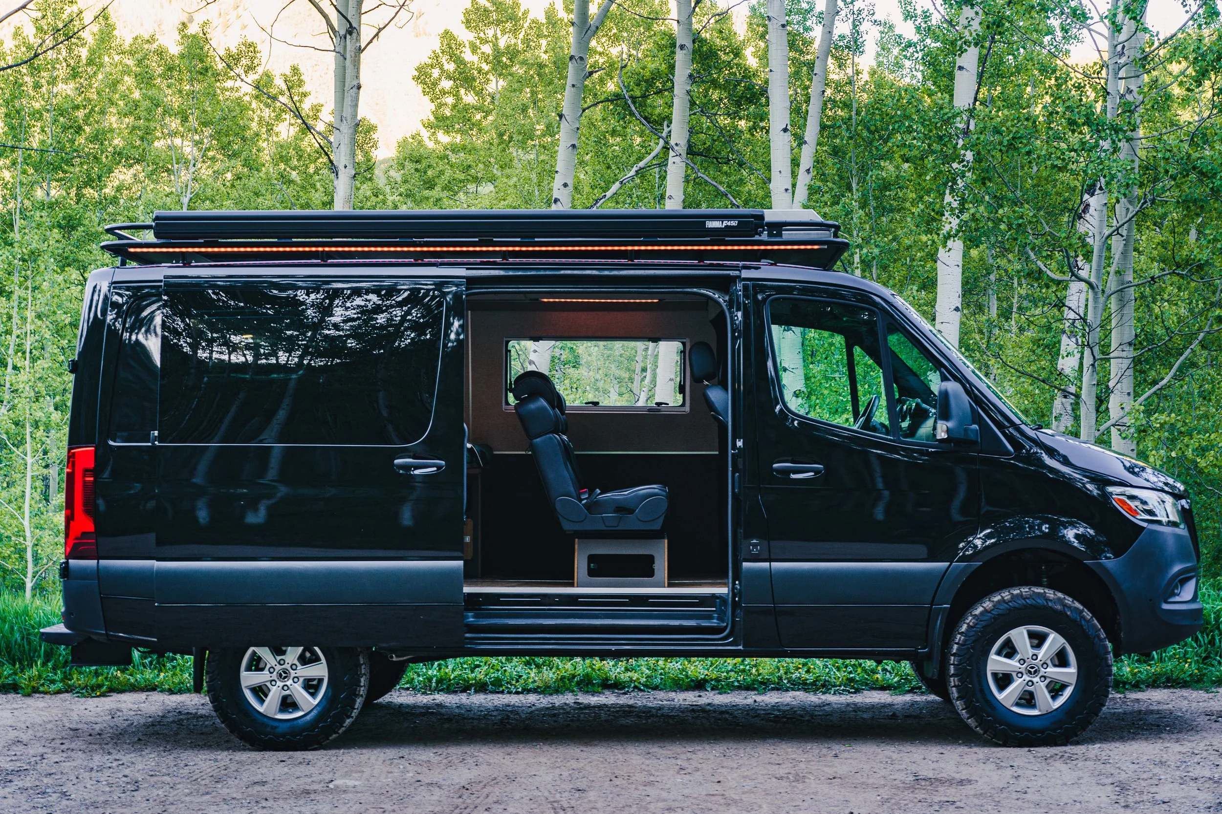 A black camper van parked in a forest with trees in the background. The van has an open sliding side door showing interior seats and a window with reflections of the trees. There is a roof rack on top.
