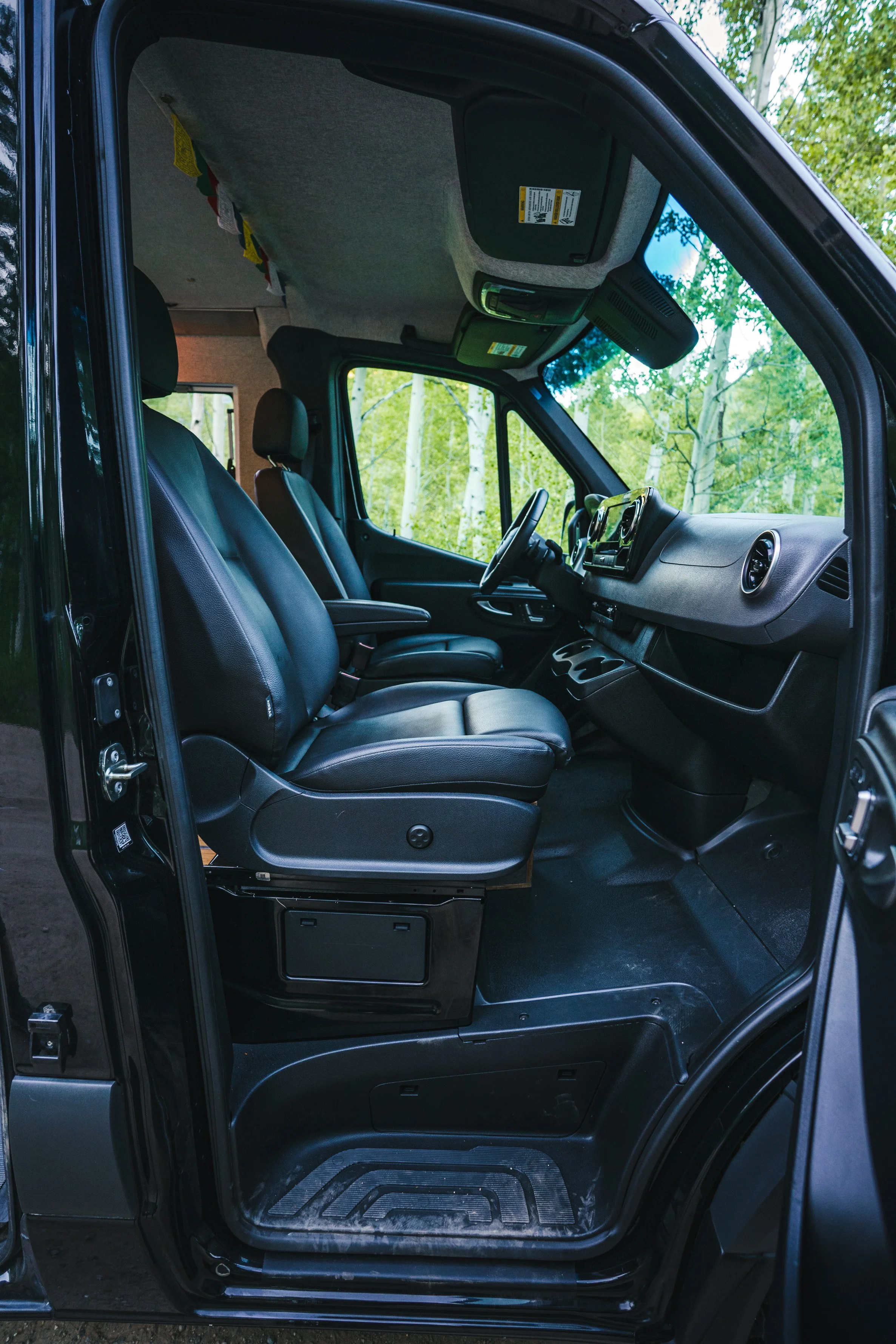 Interior of a black van showing the driver and passenger seats with black leather upholstery, dashboard, and steering wheel, surrounded by green trees visible through the windows.