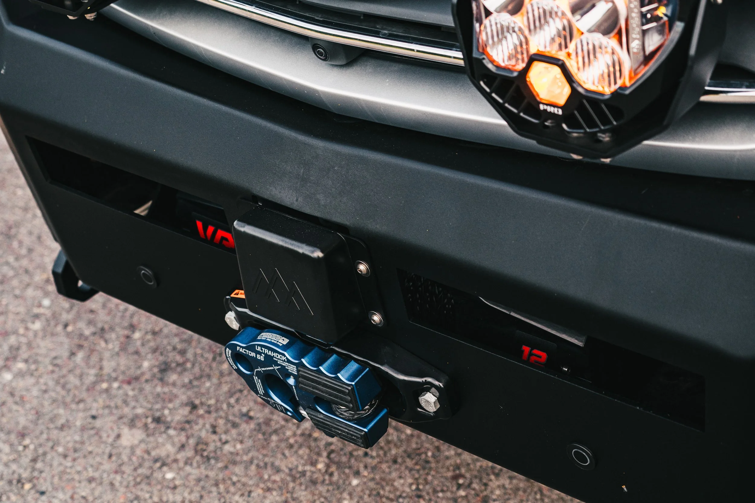 Close-up of a vehicle's front bumper showing a blue UltraHook winch, a black tow hook cover, and a license plate frame with the number 12, with a partial view of the headlight and parking sensors.