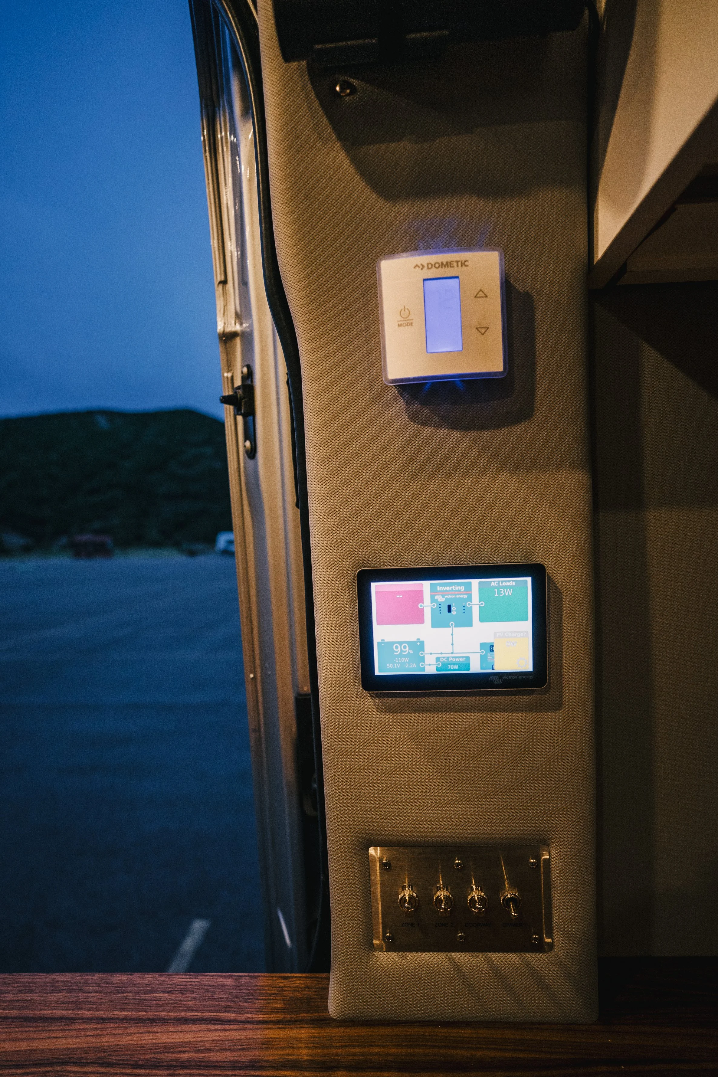 Interior of a vehicle with a control panel, including a Dometic digital controller, a digital display showing electrical system information, and switches, with an outdoor view of a parking lot and hills during dusk.