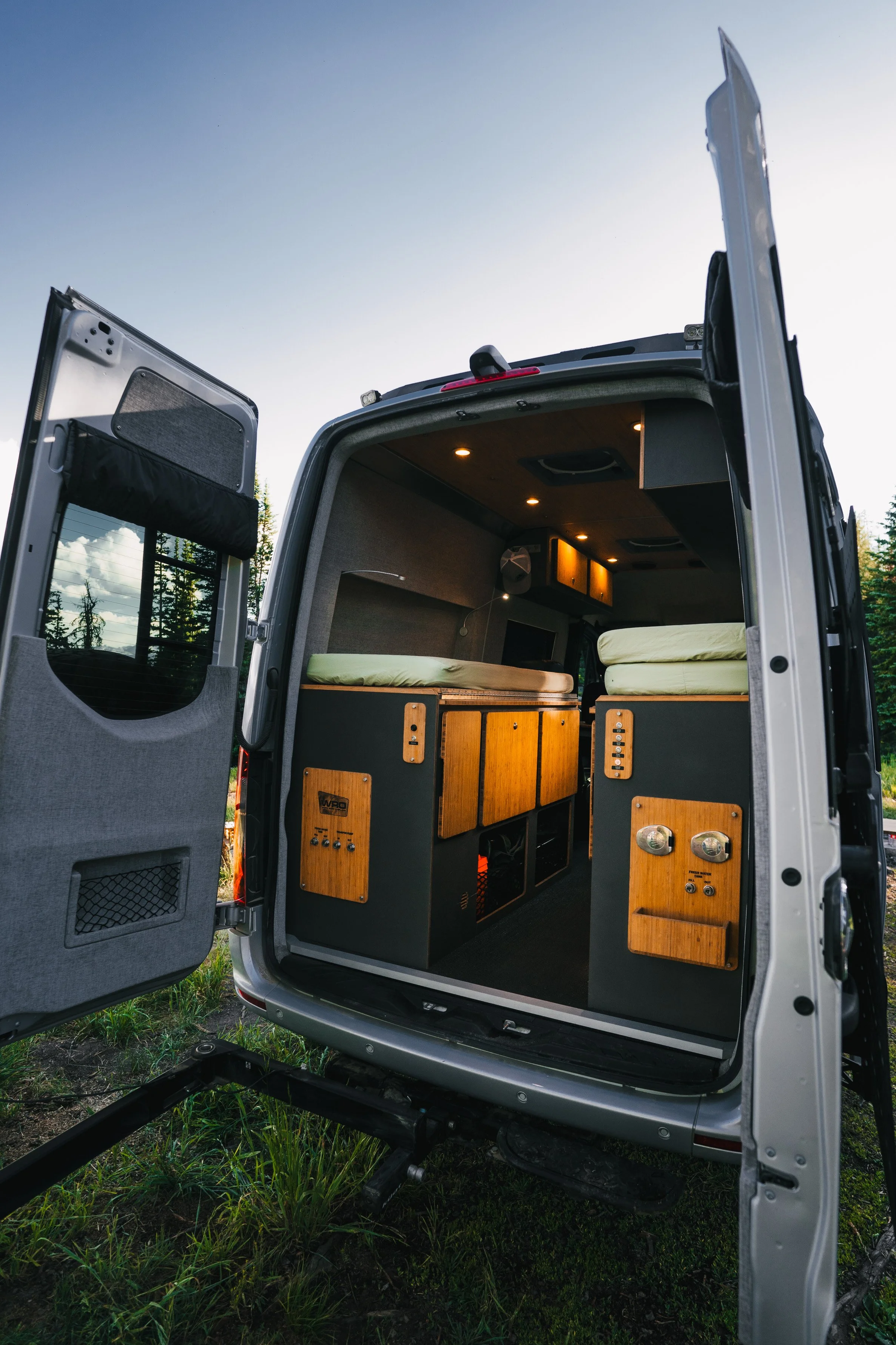 Interior of a camper van with beds, cabinets, and control panels, viewed from the open rear doors outside in a natural setting.