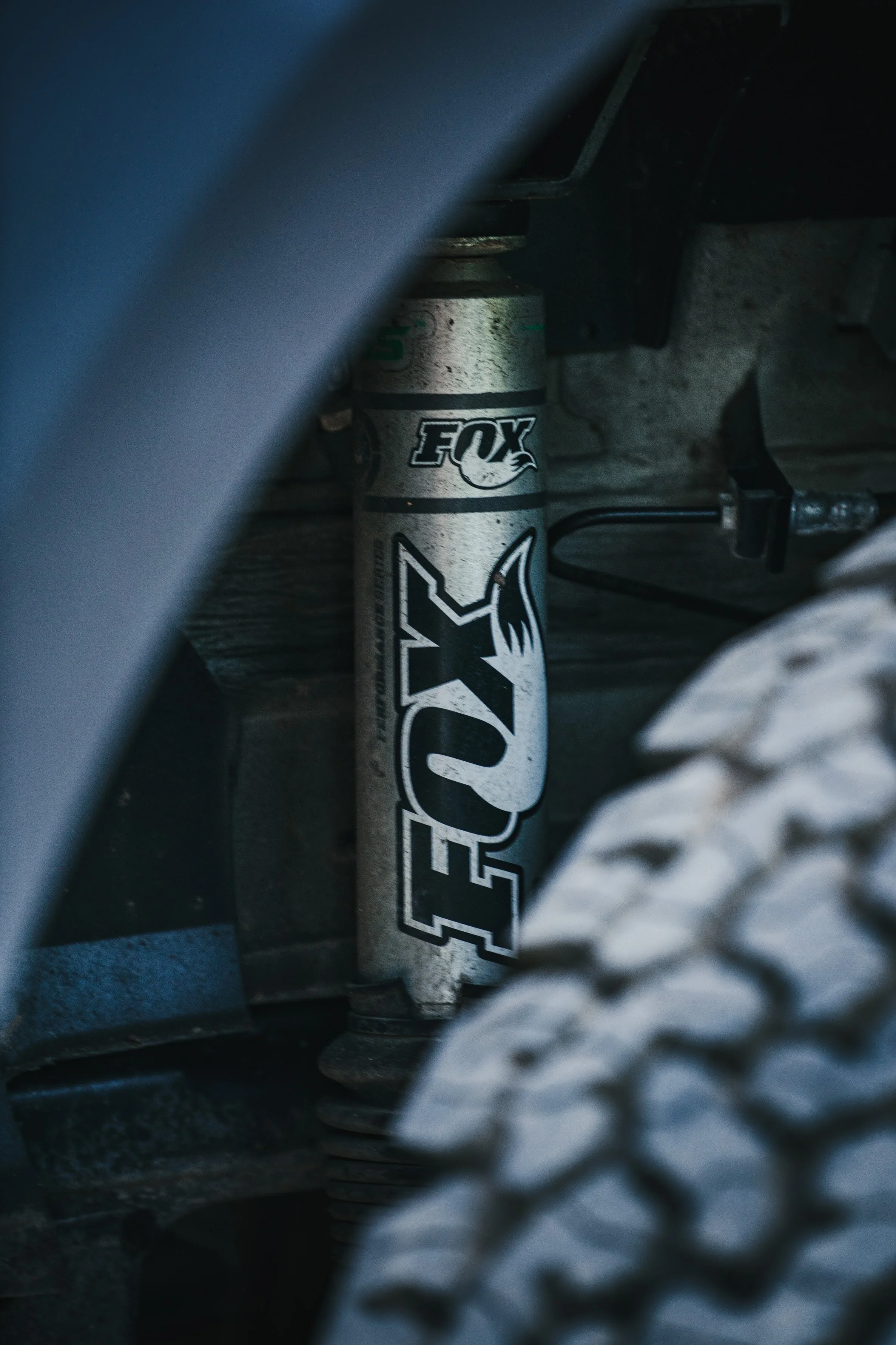 Close-up of a Fox shock absorber on a vehicle's suspension system, partially obscured by a tire and framing.