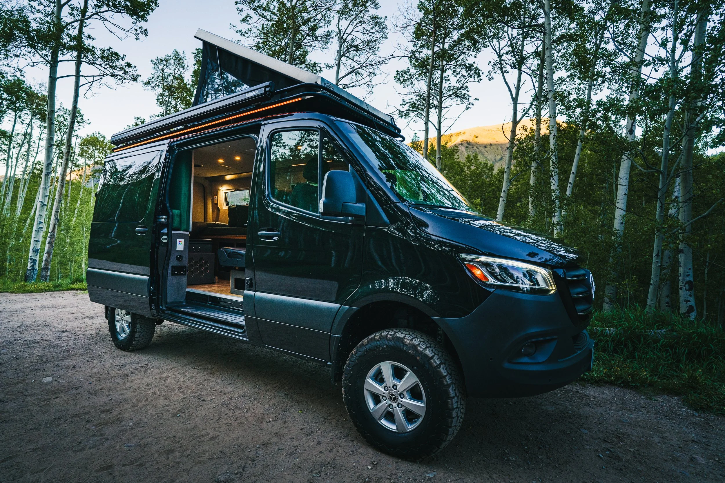 Black camper van parked on a dirt road in a forested area with trees and mountains in the background