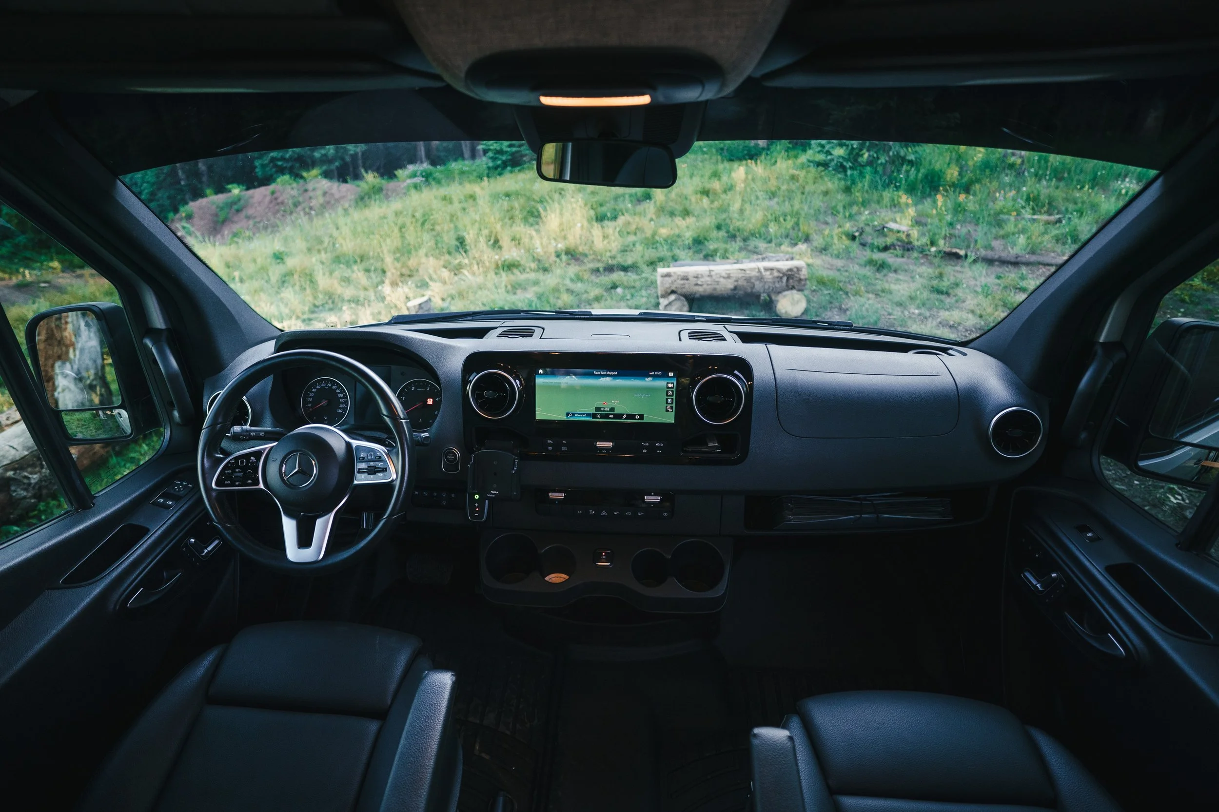 Interior view of a Mercedes-Benz van dashboard with navigation system, steering wheel, and seats, overlooking a outdoor grassy area with logs.