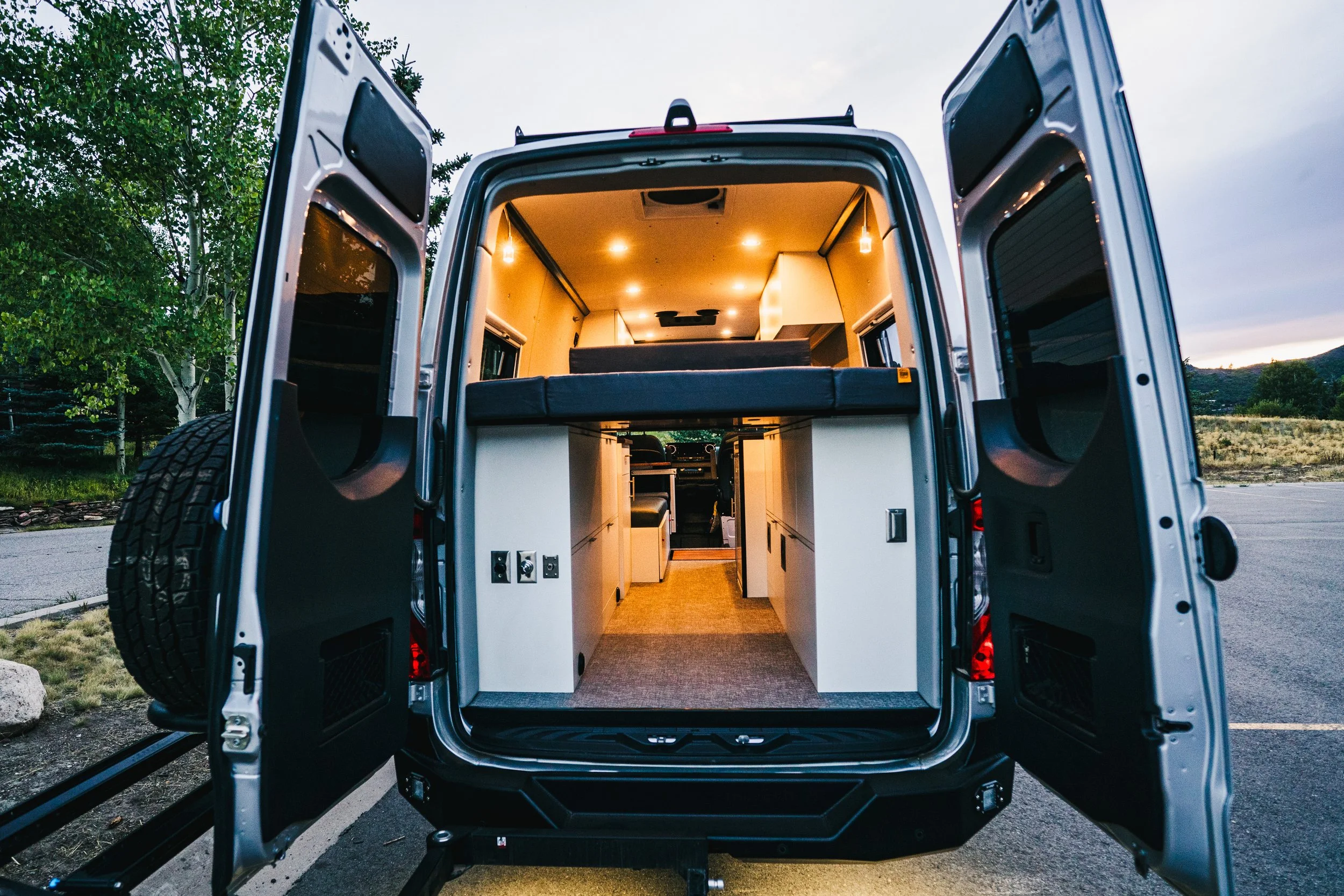The back of a mobile camper van with its doors open, showing the interior with lighting, storage cabinets, and a sleeping area, parked outdoors near trees and mountains.