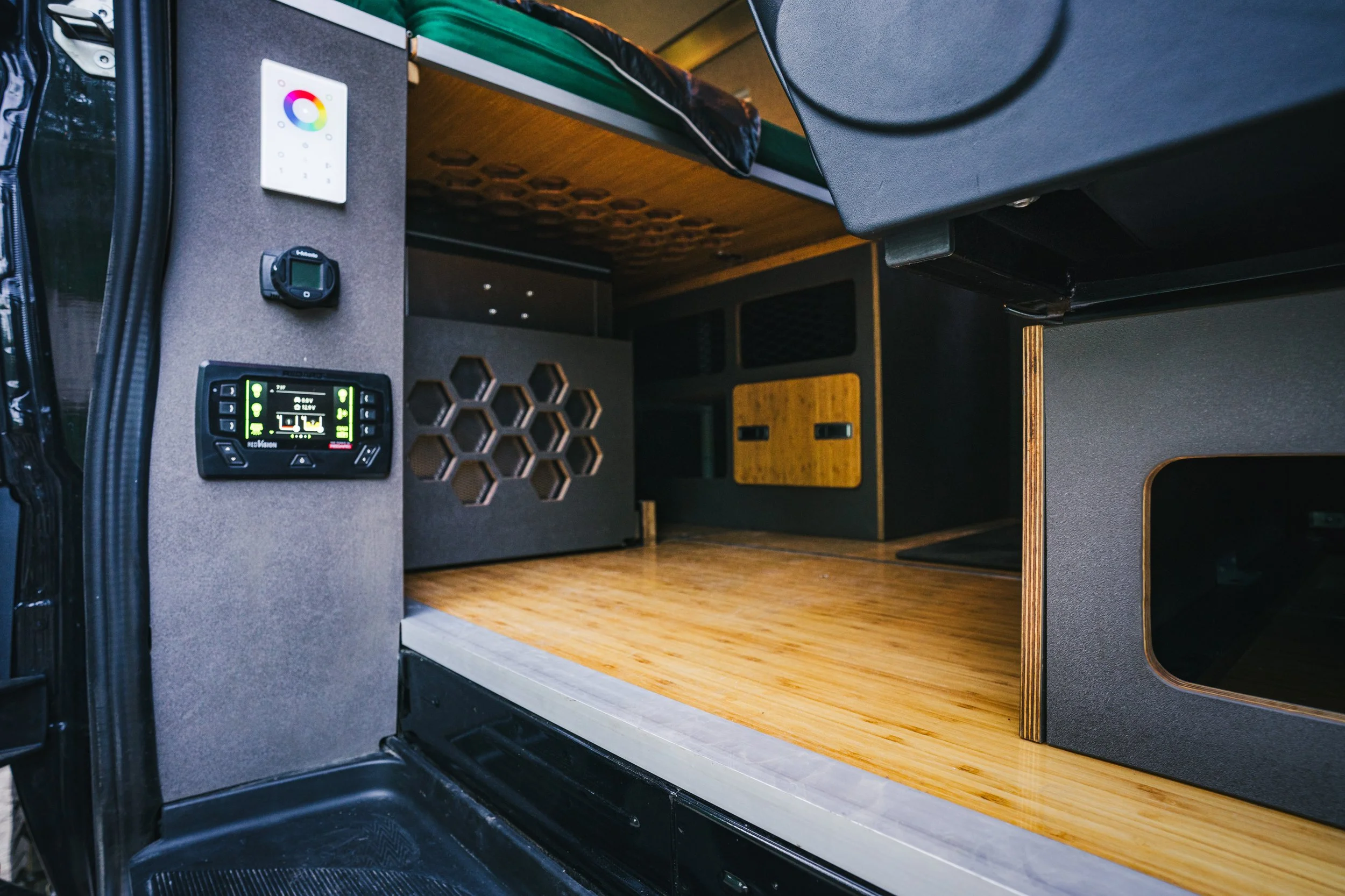 Interior of a camper van with wooden flooring, black and yellow cabinets, electronic control panel, small black device, and honeycomb wall vent.