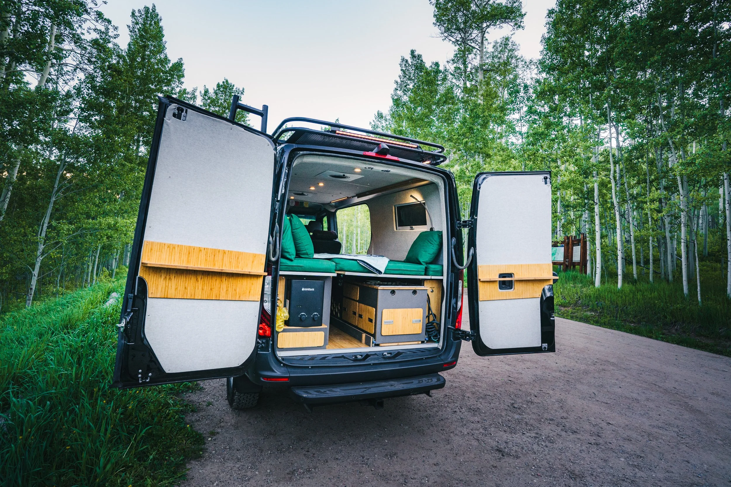 The back of a camper van parked on a dirt road in a forest, with open doors revealing a cozy interior with green cushions and wood accents.