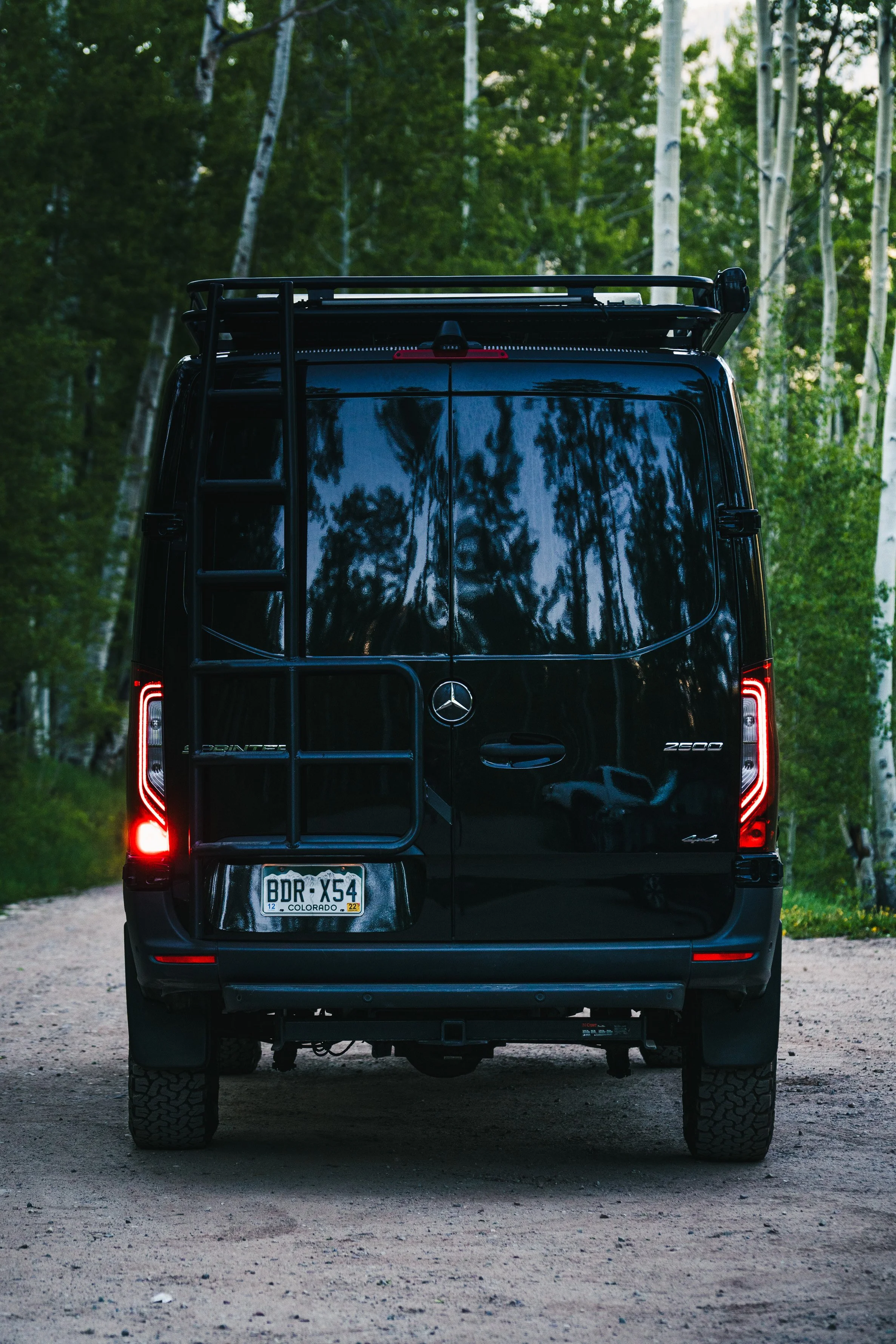 Rear view of a black Mercedes-Benz Sprinter van with Colorado license plate, on a dirt road in a forested area with tall trees.