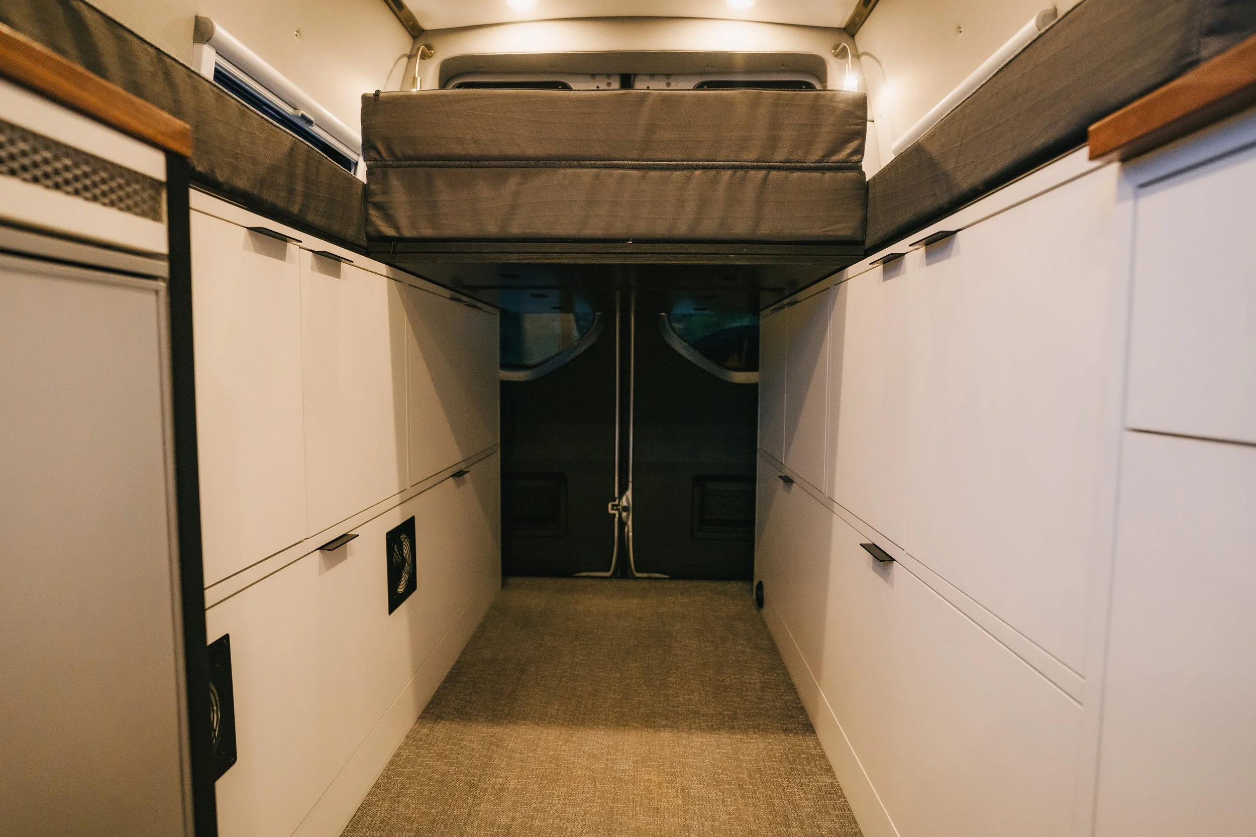 View of a small, modern camper van interior with white cabinets on both sides, a carpeted floor, and a sleeping loft with a brown mattress and a brown storage container underneath.