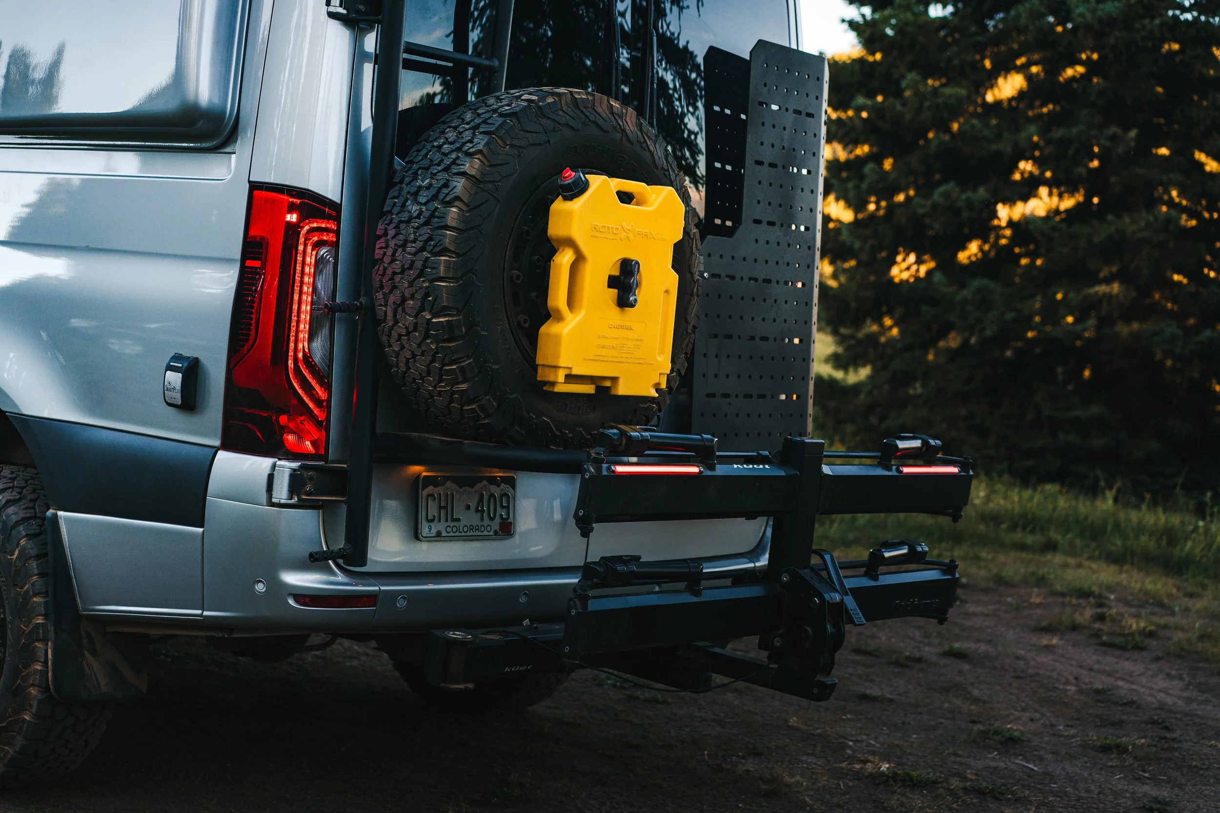 Rear view of a silver off-road vehicle with a mounted spare tire, a yellow fuel container, and a black bike rack, parked on a dirt surface with trees in the background at sunset.