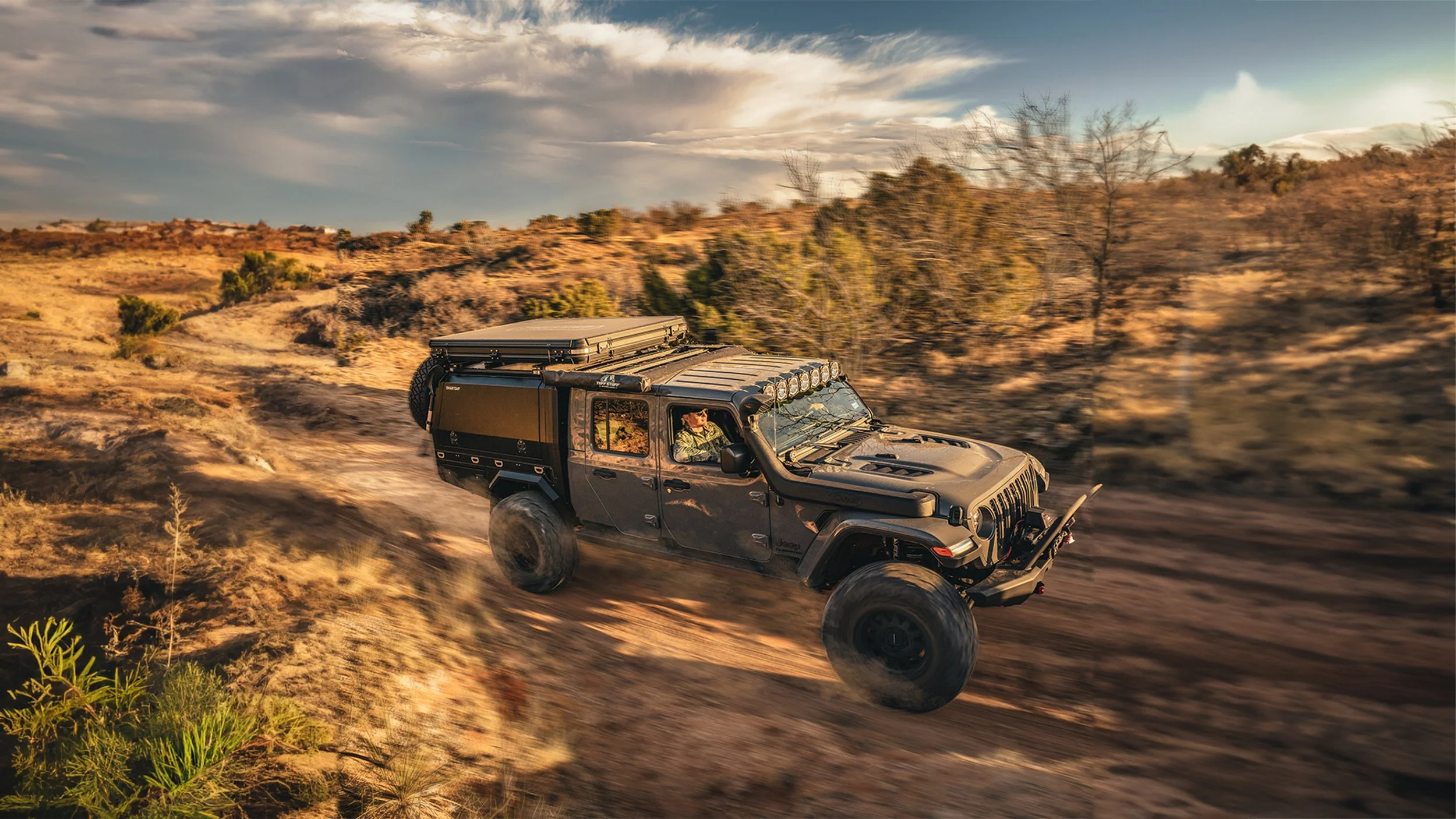 Black off-road vehicle driving on a dirt trail through a semi-arid landscape with sparse shrubs and trees, under a sky with scattered clouds during sunset.