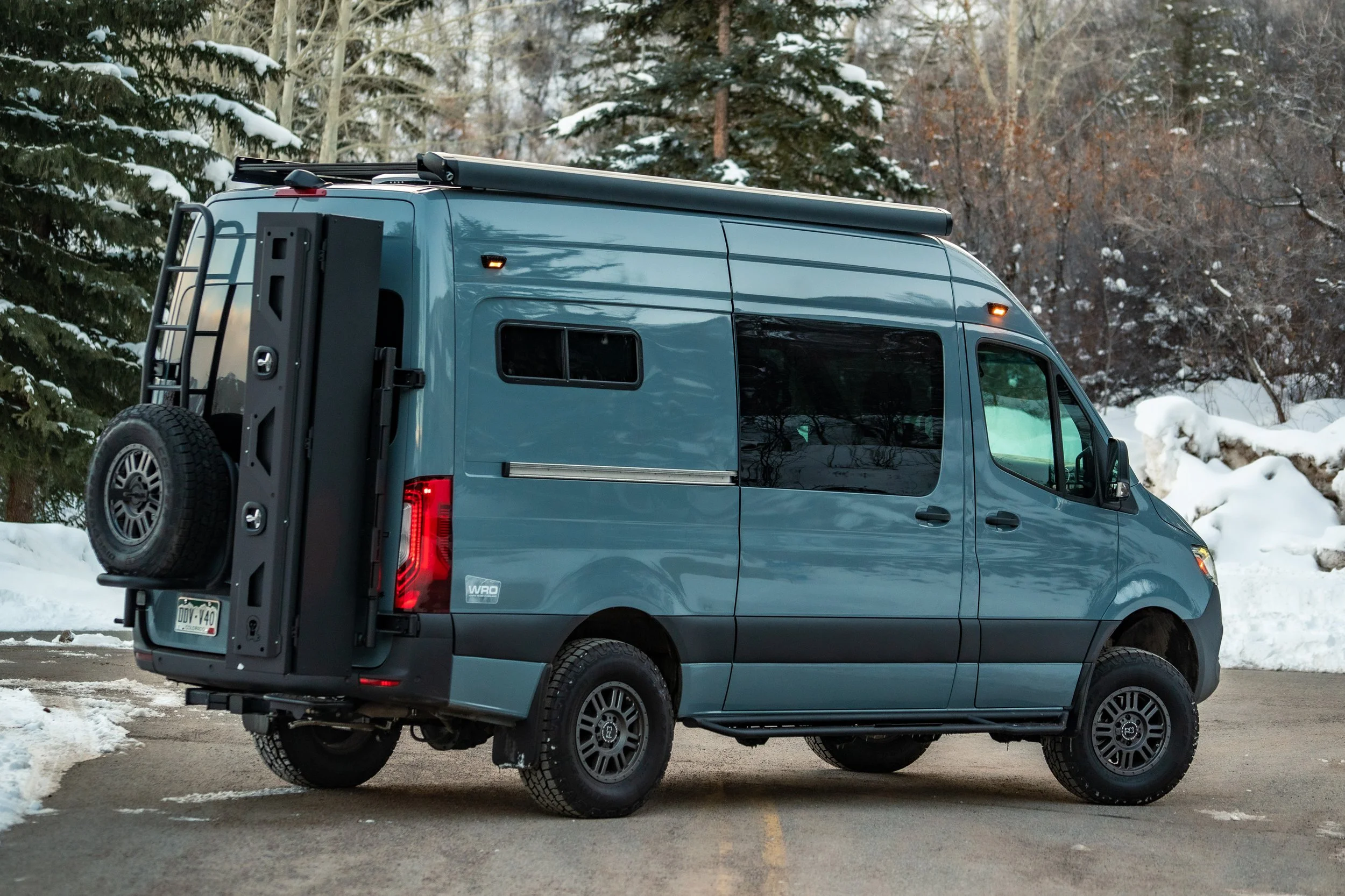 A blue camper van parked in a snowy forested area, featuring a spare tire mounted on the back, black wheels, and a roof awning.