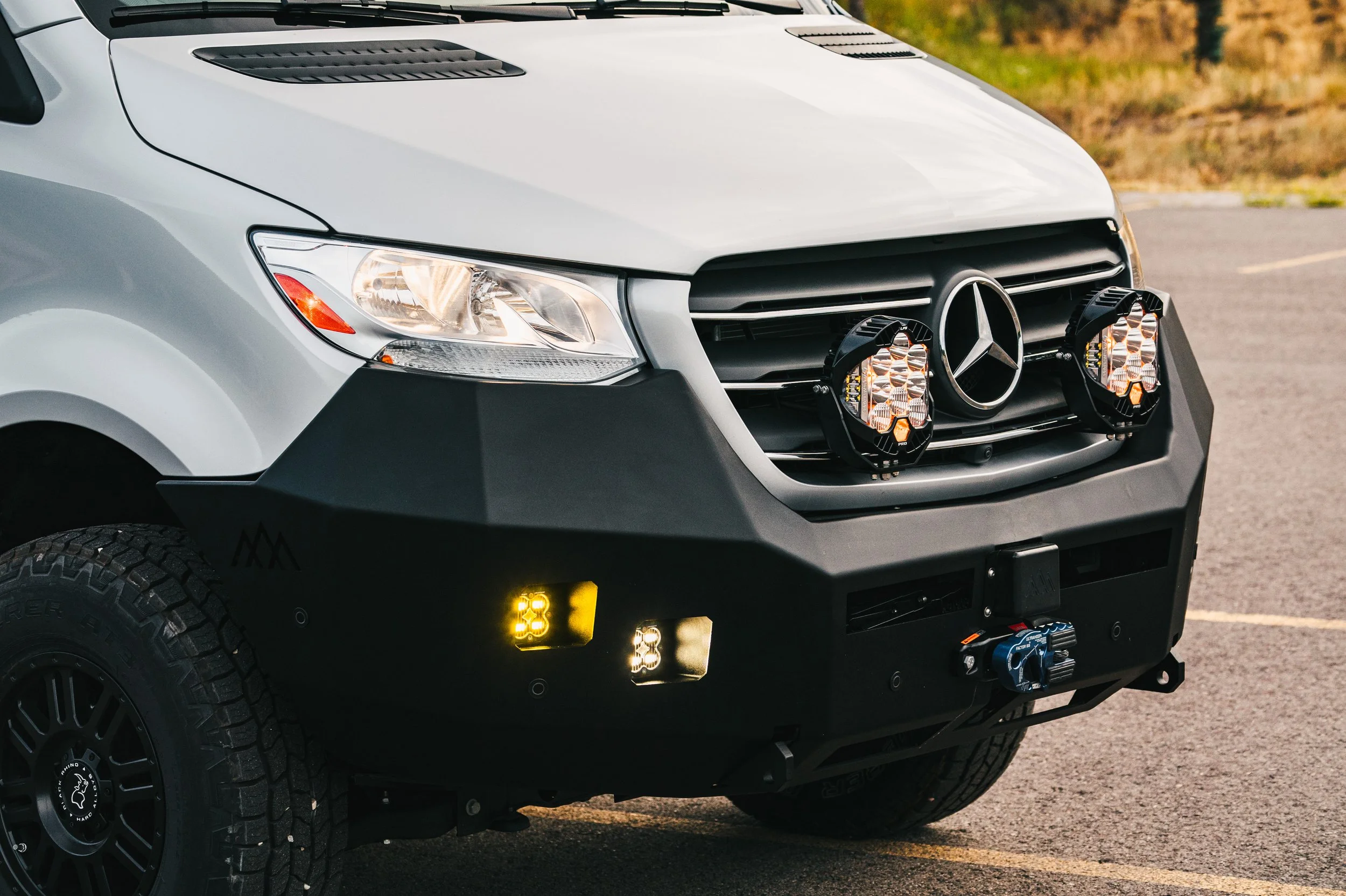 Close-up of a silver Mercedes-Benz Sprinter van equipped with off-road modifications, including additional lights on the front grille, a heavy-duty bumper, and off-road tires, parked in an outdoor lot.