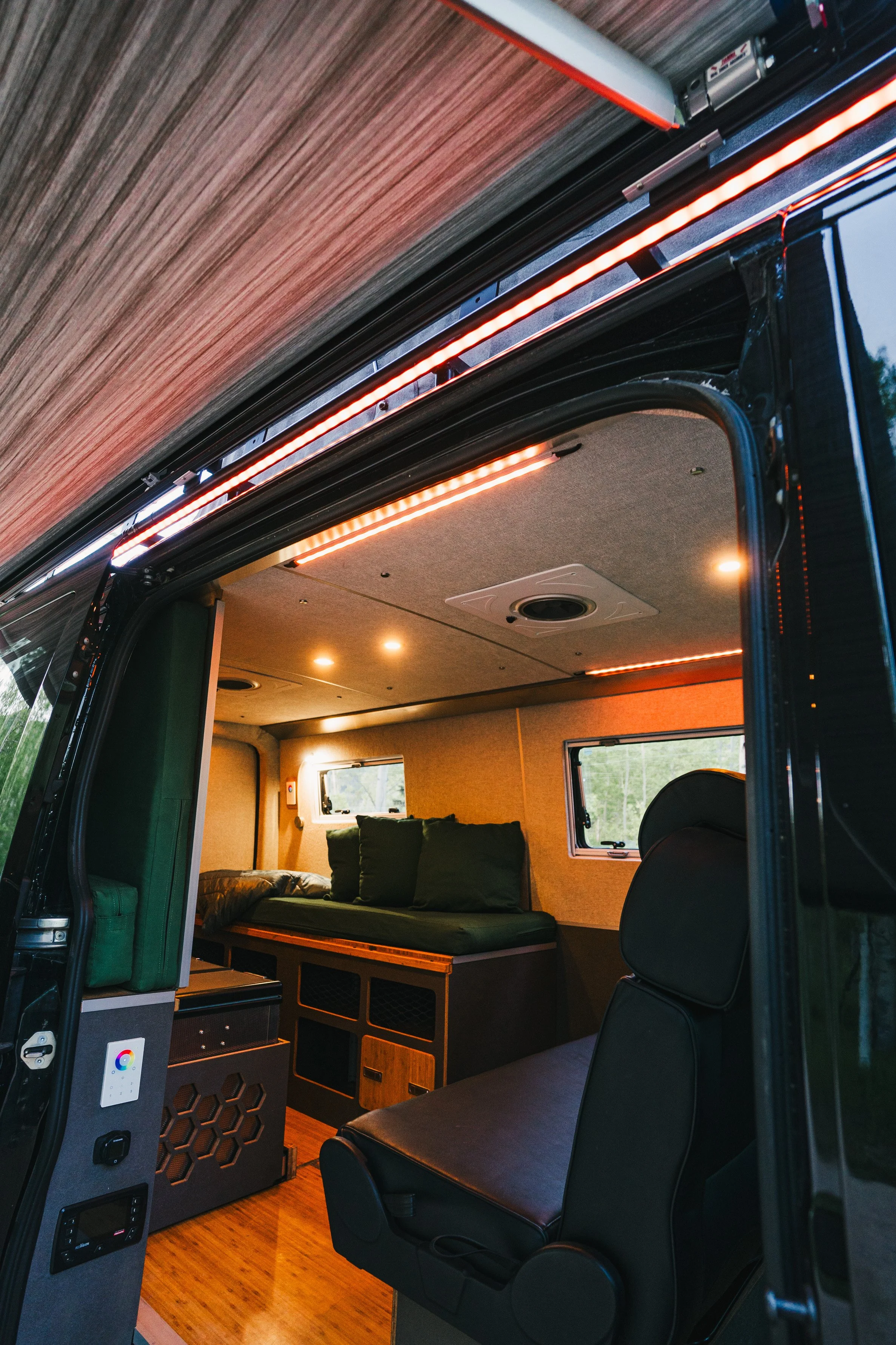 Interior of a modern camper van showing a seating area with window, cushioned bench, and wood accents, illuminated by warm LED strip lights.