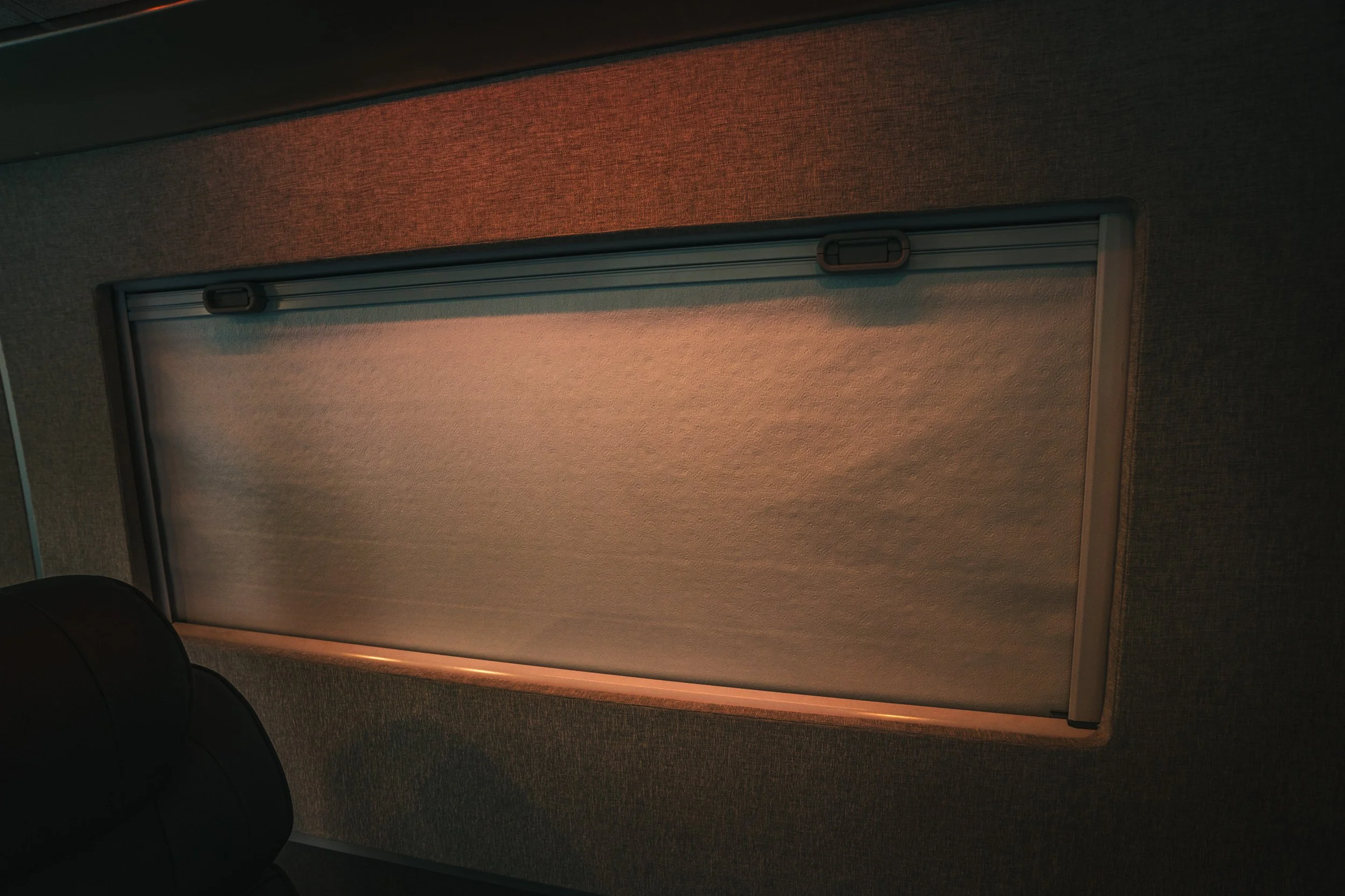 An interior window with a closed shade or blind, framed by textured dark gray wall, and a black leather seat in the lower corner.