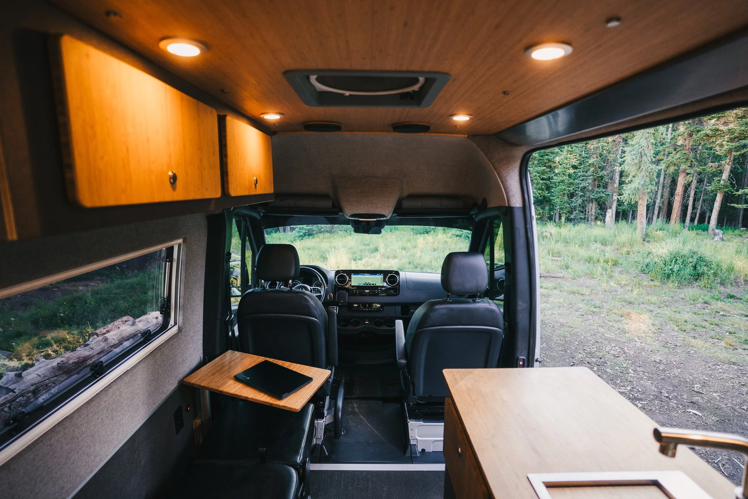 The interior of a camper van showing the driver's and passenger's seats, a small wooden table with a closed black notebook, overhead wooden cabinets, and a window view of a grassy area with trees.