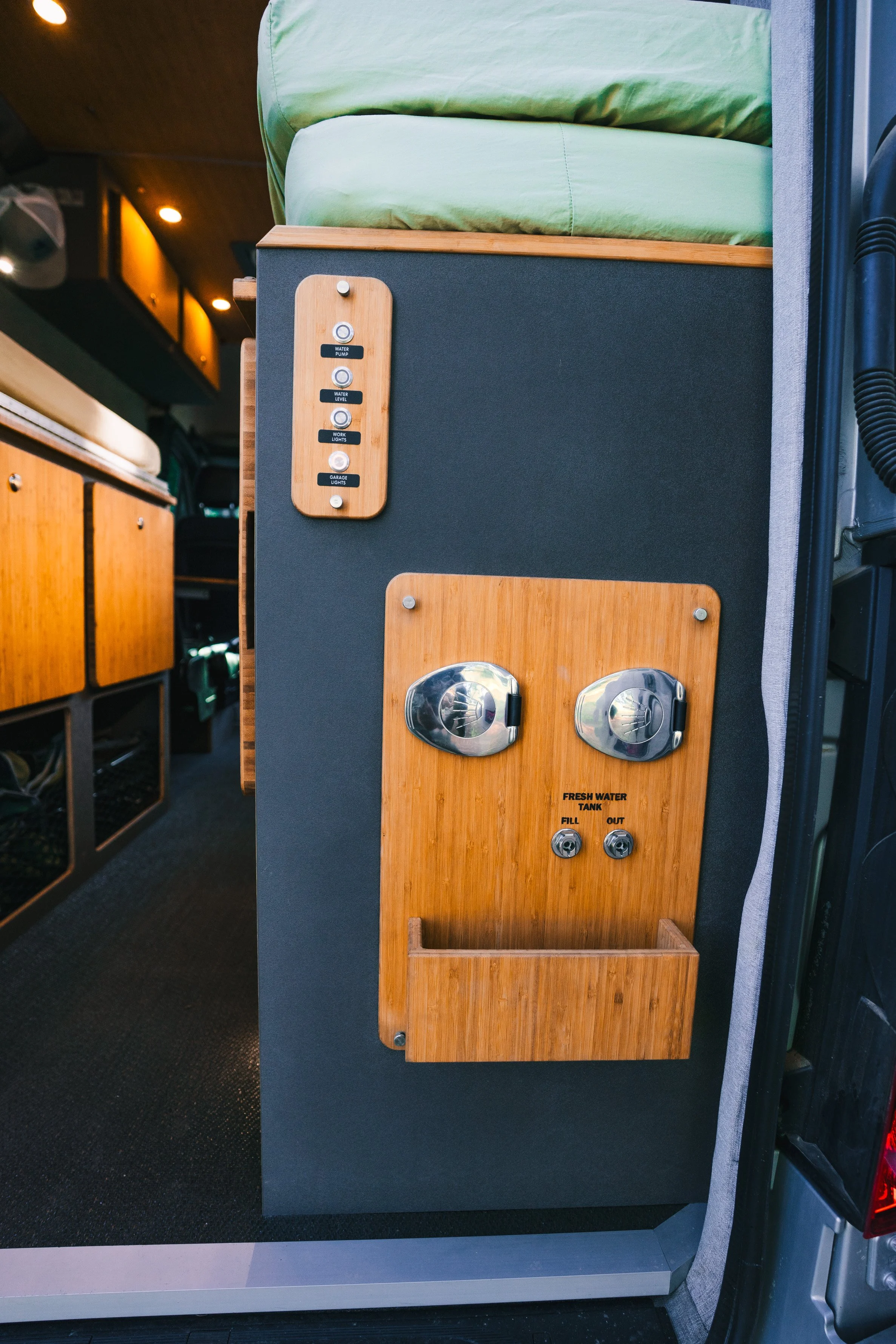 Interior of a camper van showing a side panel with control switches, a fresh water tank with fill and out ports, and some green bedding on top.