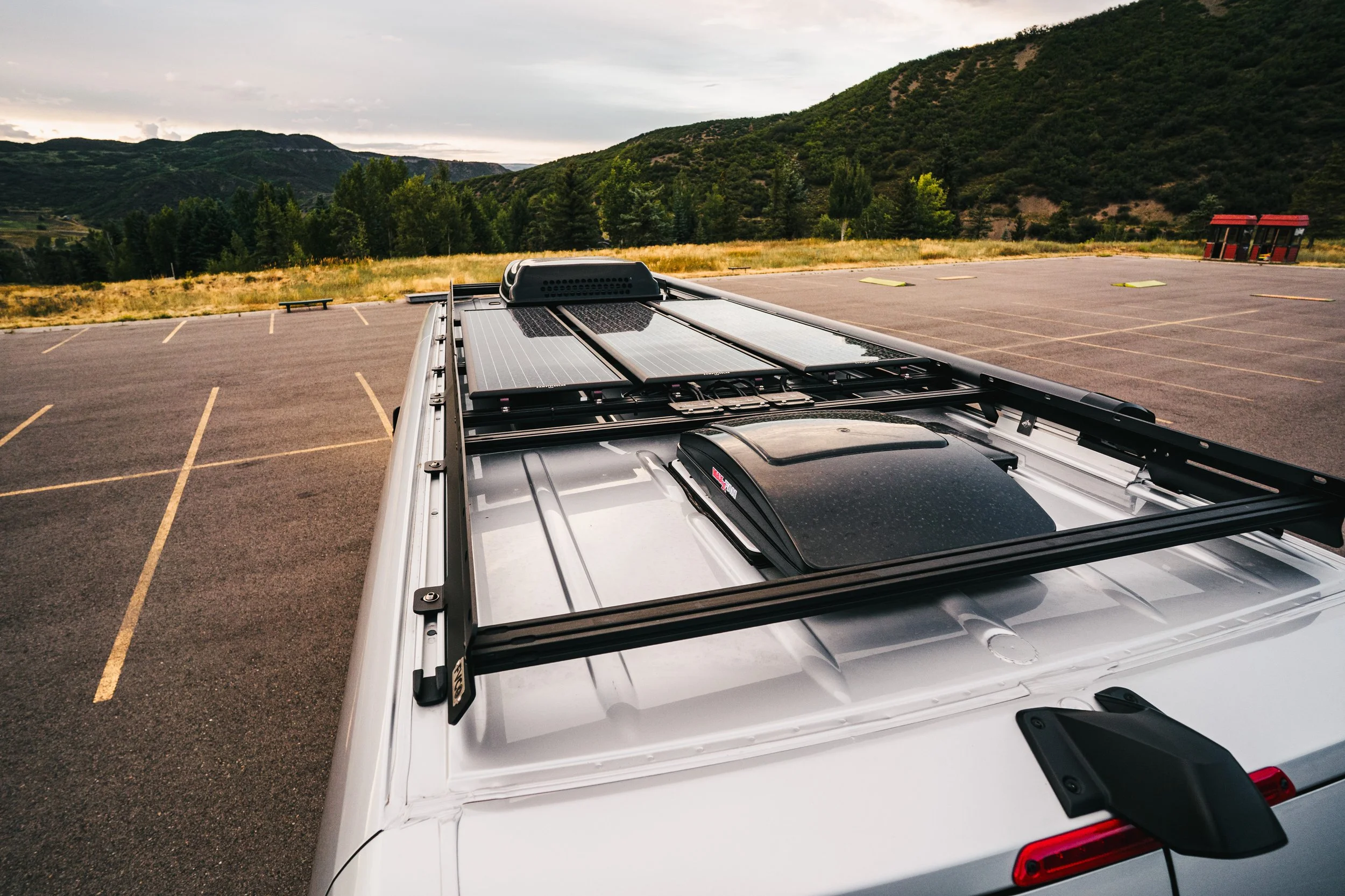 The roof of a Sprinter van equipped with solar panels and storage compartments, parked in an empty lot with hills and mountains in the background.