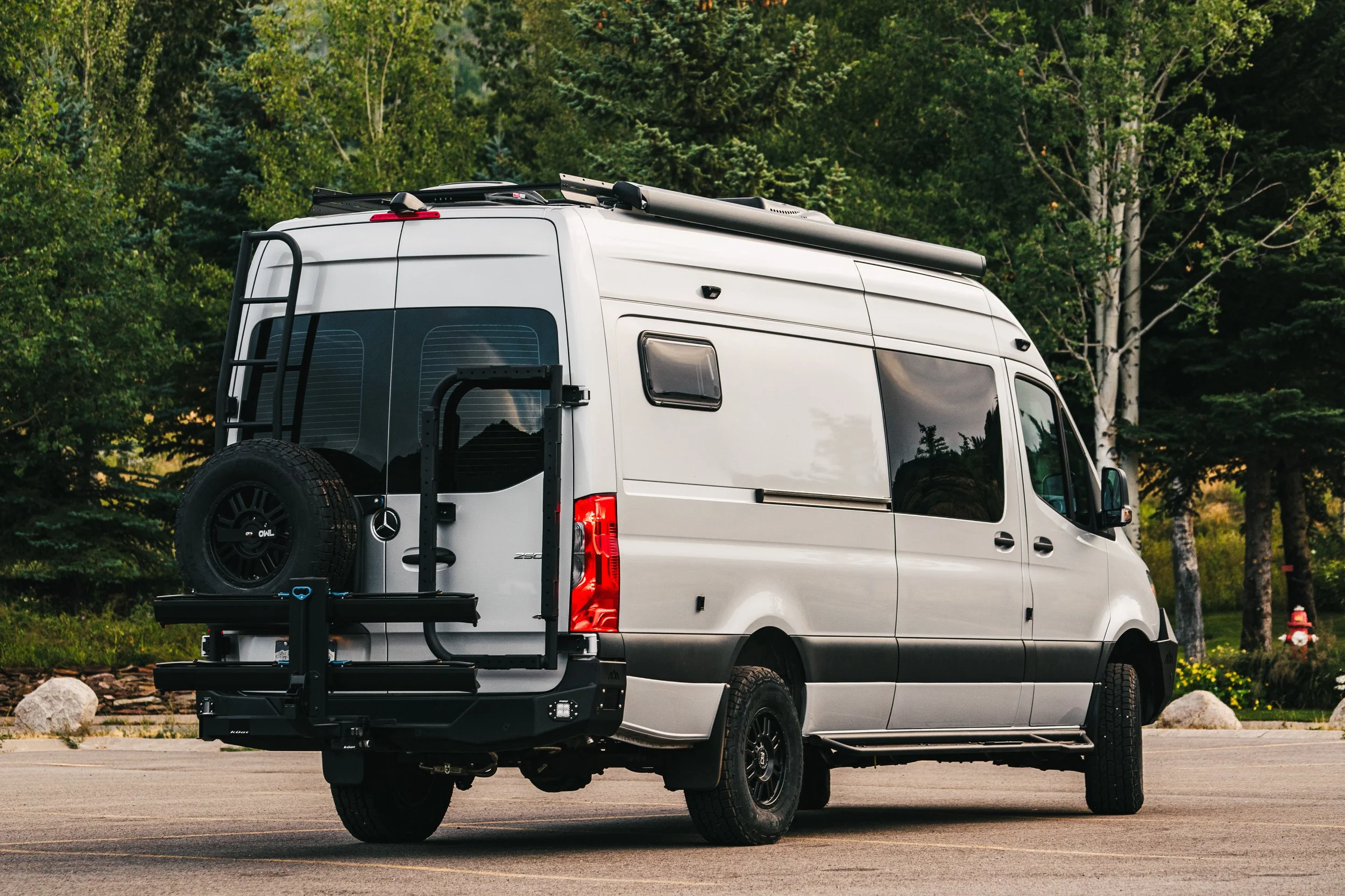 A silver Sprinter camper van with a spare tire mounted on the back, parked on a paved surface with trees in the background.