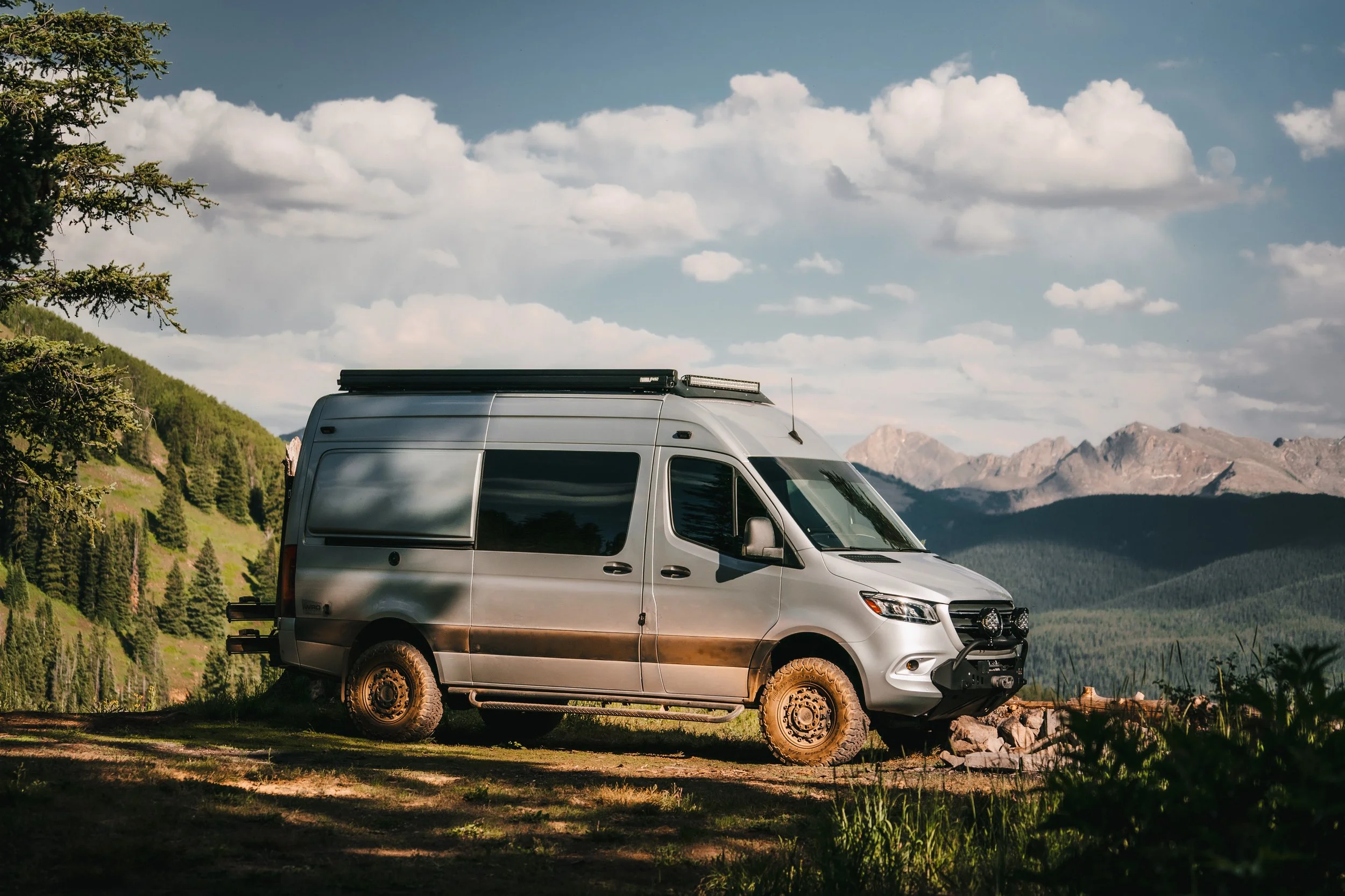 Silver camper van parked in a mountainous landscape with trees, clouds, and blue sky in the background.