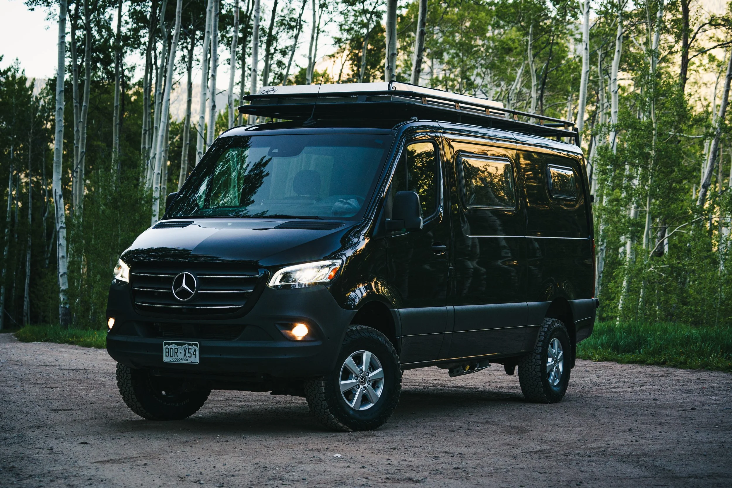 Black Mercedes-Benz Sprinter van parked on a dirt road in a forest with trees and green foliage, during sunset.