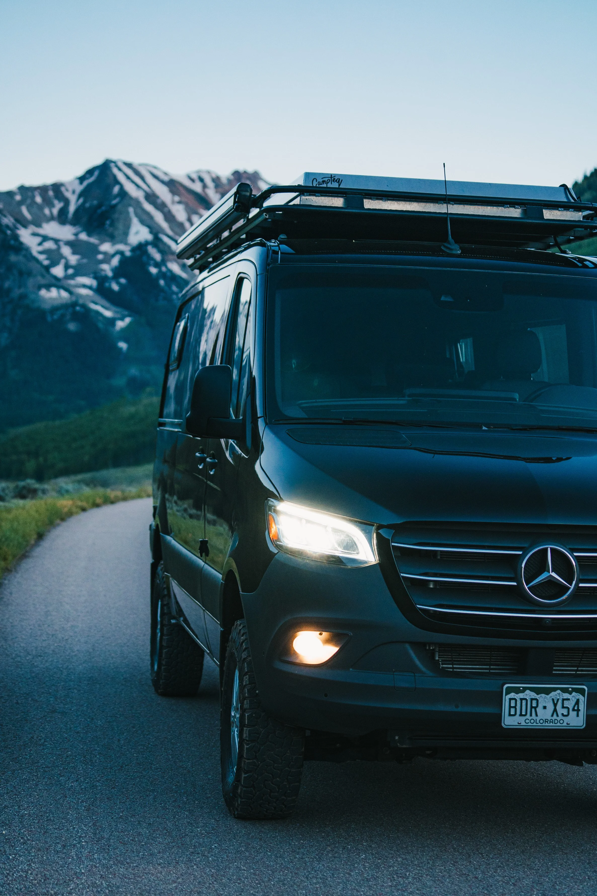 A black Mercedes-Benz camper van parked on a mountain road with snow-capped peaks in the background, during dusk or dawn.