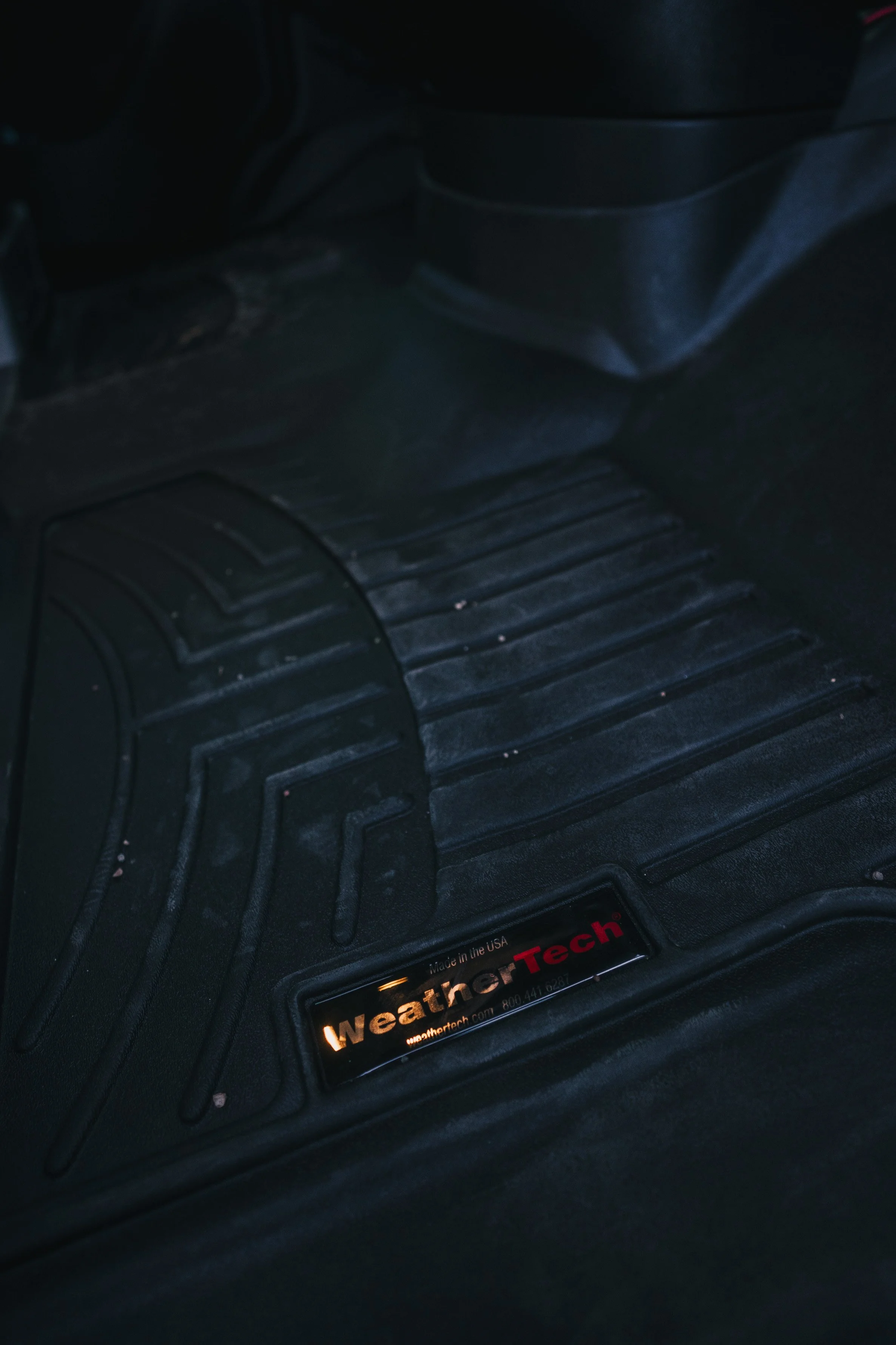 Close-up of a black WeatherTech car floor mat with a red and white WeatherTech label, showing some dirt and debris.