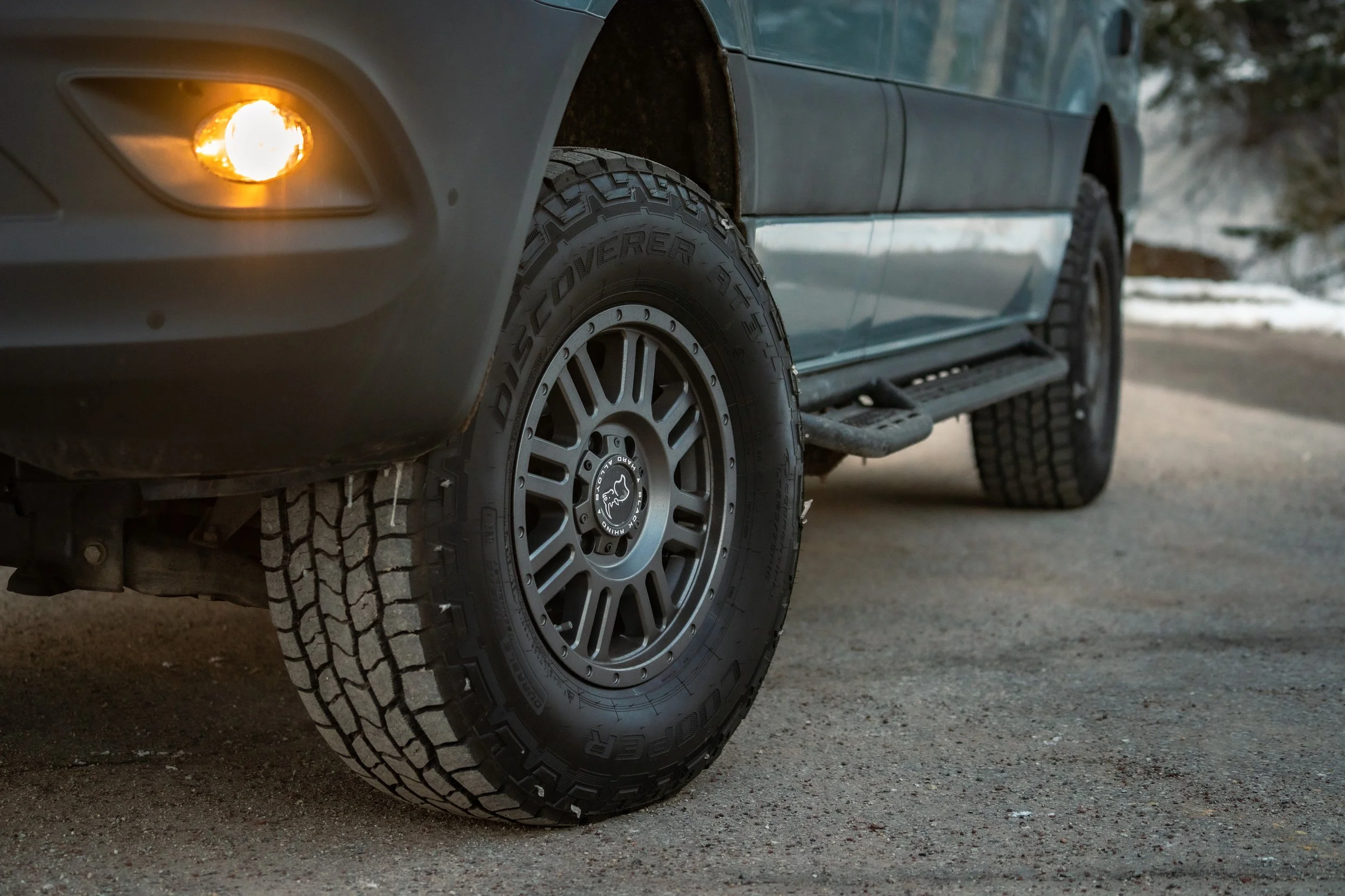 Close-up of the front wheel and tire of a dark-colored SUV parked on a gravel surface, with a snowy landscape in the background.