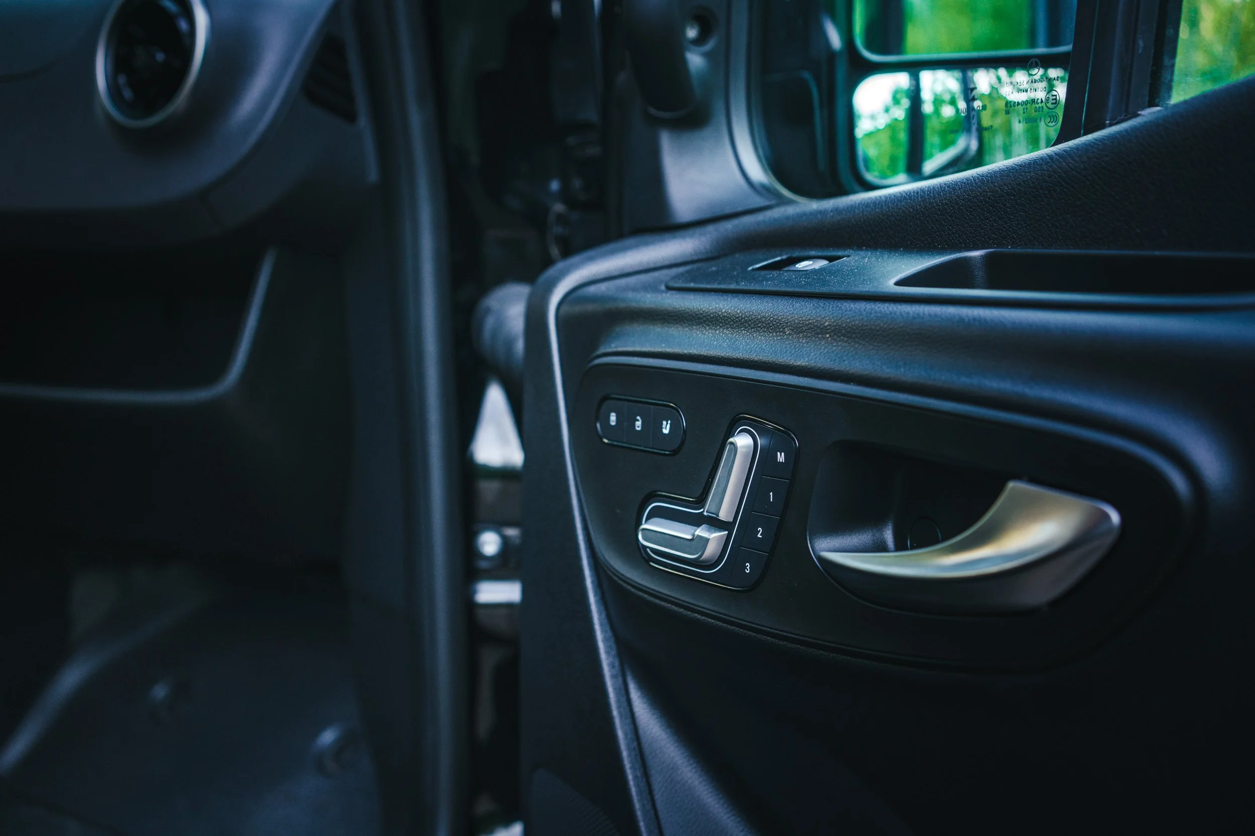 Close-up of a car door panel with seat adjustment controls, including buttons labeled M, 1, 2, and 3, and a metallic door handle.